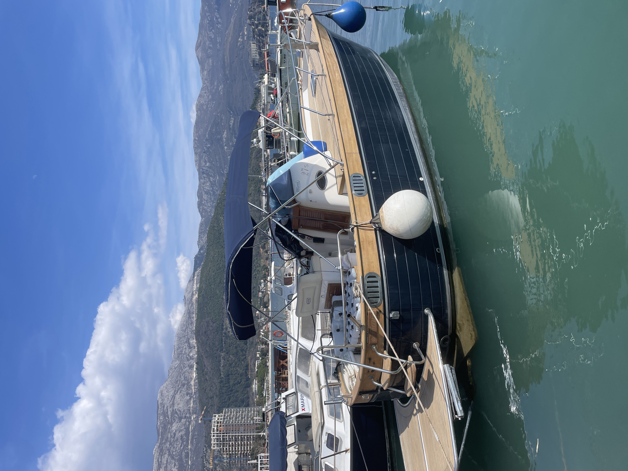 The image displays a marina in Budva, Montenegro, under a clear blue sky with scattered white cumulus clouds. A dark blue motor yacht, labeled "Mimi 27s," is moored in the foreground to the midground, oriented with its bow facing right. This yacht features a light wooden deck, stainless steel railings, a wooden swim platform with integrated steps at the stern, and a blue canvas bimini top covering the cockpit area. A white spherical fender is attached to its port side, and a blue spherical fender is attached to its starboard bow. Other vessels are partially visible to the left and in the background, including a white motorboat with "XMARINA" displayed on its hull and another boat with "17-BD." The background comprises a range of rugged, grey-brown mountains. Situated at the base of these mountains is a section of Budva city, where a multi-story building under construction with a visible crane is prominent on the left, alongside other coastal structures. The calm, turquoise water reflects the boats and sky. No individuals are visible in the scene.