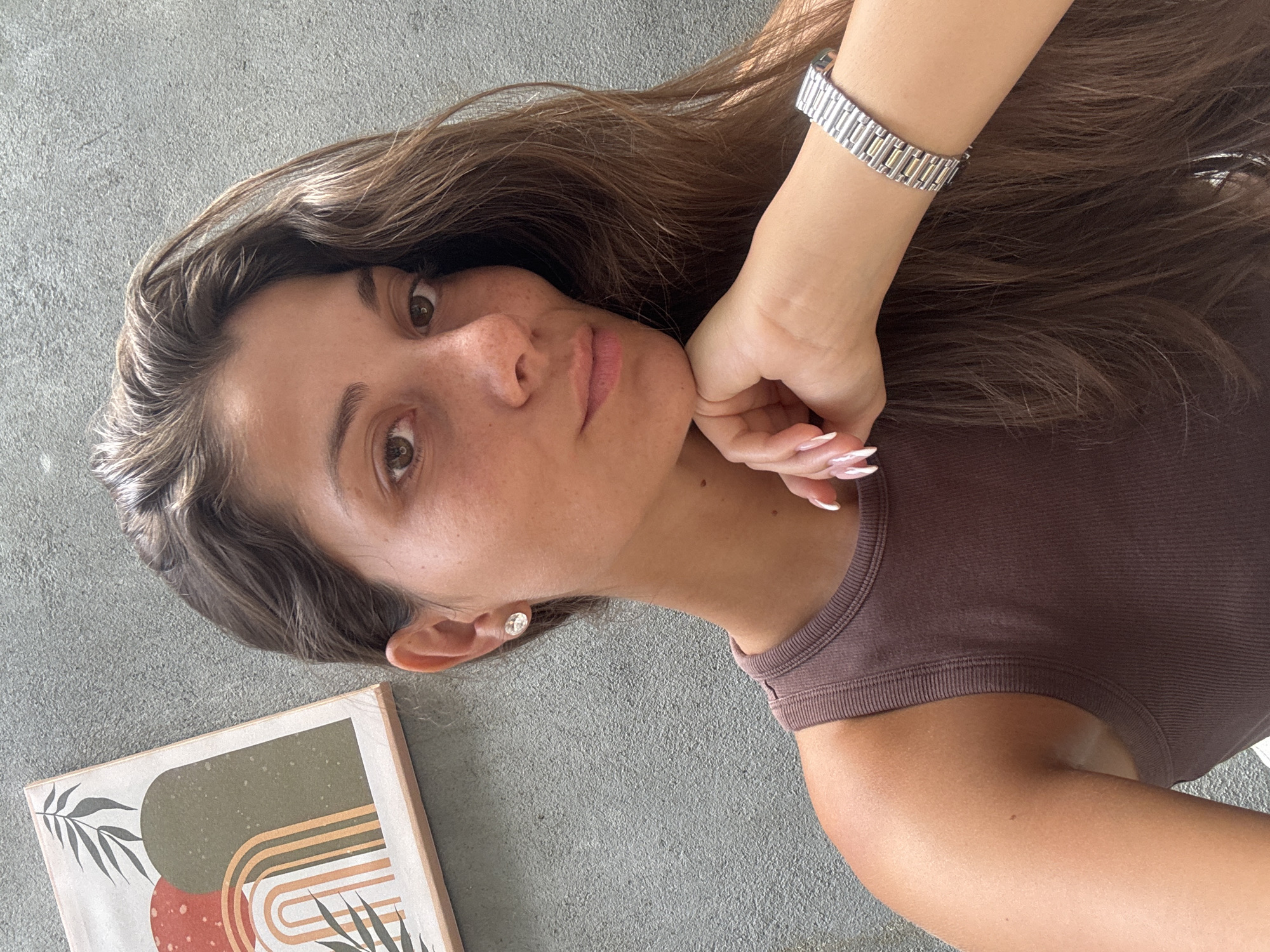 A medium close-up portrait shows a woman lying on a gray concrete surface. She has light brown skin, freckles on her face, and brown eyes. Her long, wavy brown hair is spread around her head and shoulders. She is wearing a brown tank top and a silver bracelet on her left wrist. Her left hand is positioned near her chin, with manicured nails. To the lower left of the frame is a framed picture with abstract shapes and colors. No specific location in Đà Nẵng, Vietnam, can be determined from the image.