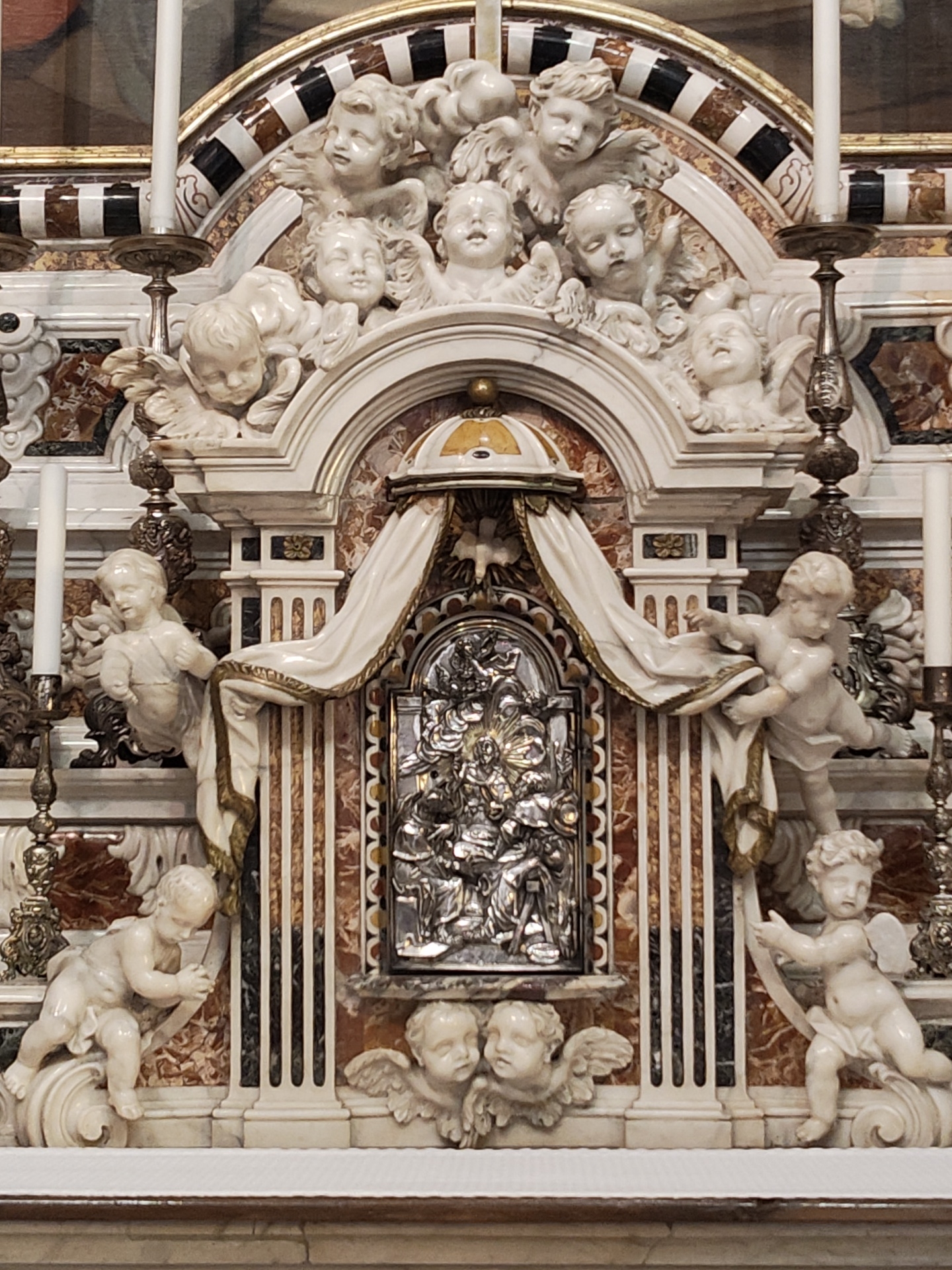 A highly ornate altarpiece, likely located in a church in Cagliari, Italy, is depicted. The altarpiece is constructed of marble in varying shades of brown, white, and black, and features carved white cherubs throughout its design. At the center of the altarpiece is a silver relief, likely a depiction of a religious scene. A white draped cloth hangs above the relief. On either side of the relief are white marble pillars with carved floral accents. Carved cherubs cling to these pillars. Above the pillars, more cherubs are carved in the marble, filling the upper arch of the structure. Atop the arch is a dome-like structure, featuring a golden top. Three white candles in ornate silver candleholders flank the upper arch. At the base of the structure are two cherubic faces with wings, carved in white marble. The background of the altarpiece shows more dark brown marble with white veins.