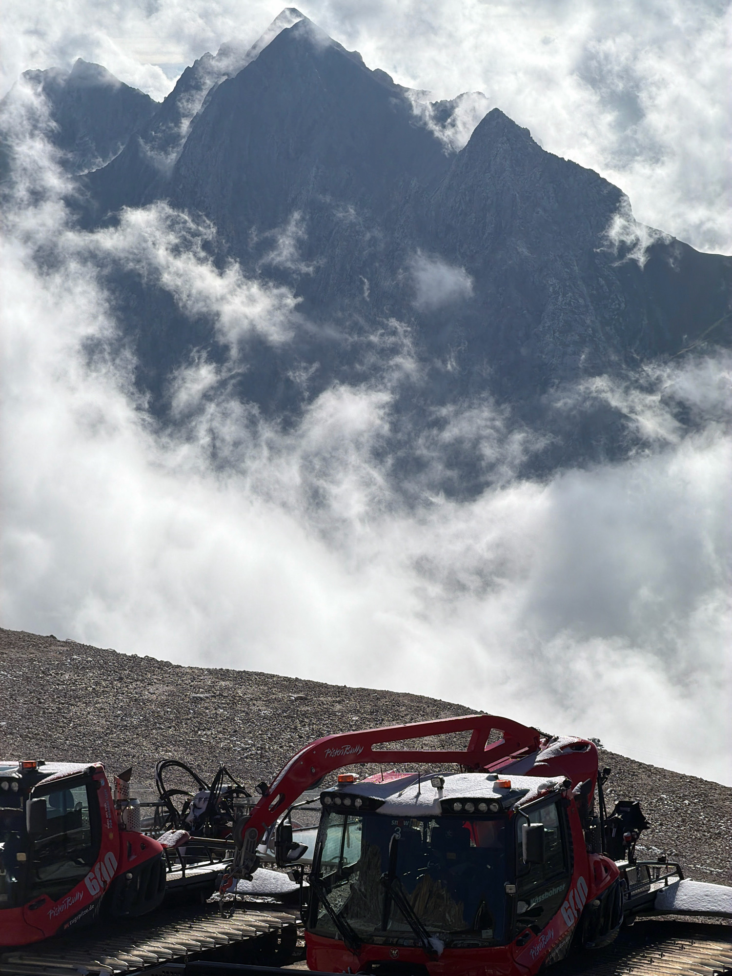The image shows a view from the Zugspitze, Germany's highest mountain, located near Garmisch-Partenkirchen. In the foreground, two red PistenBully snow groomers are parked on a gravelly slope, dusted with snow. One snow groomer on the left is partially visible, displaying the number "610" and the PistenBully logo. The snow groomer on the right is more prominent, also showing the PistenBully branding and the model number "610." It has a mechanical arm extended. The background features dramatic, jagged mountain peaks partially obscured by swirling clouds. The sky is overcast with patches of bright light filtering through. The overall scene depicts a rugged, high-altitude mountain environment.