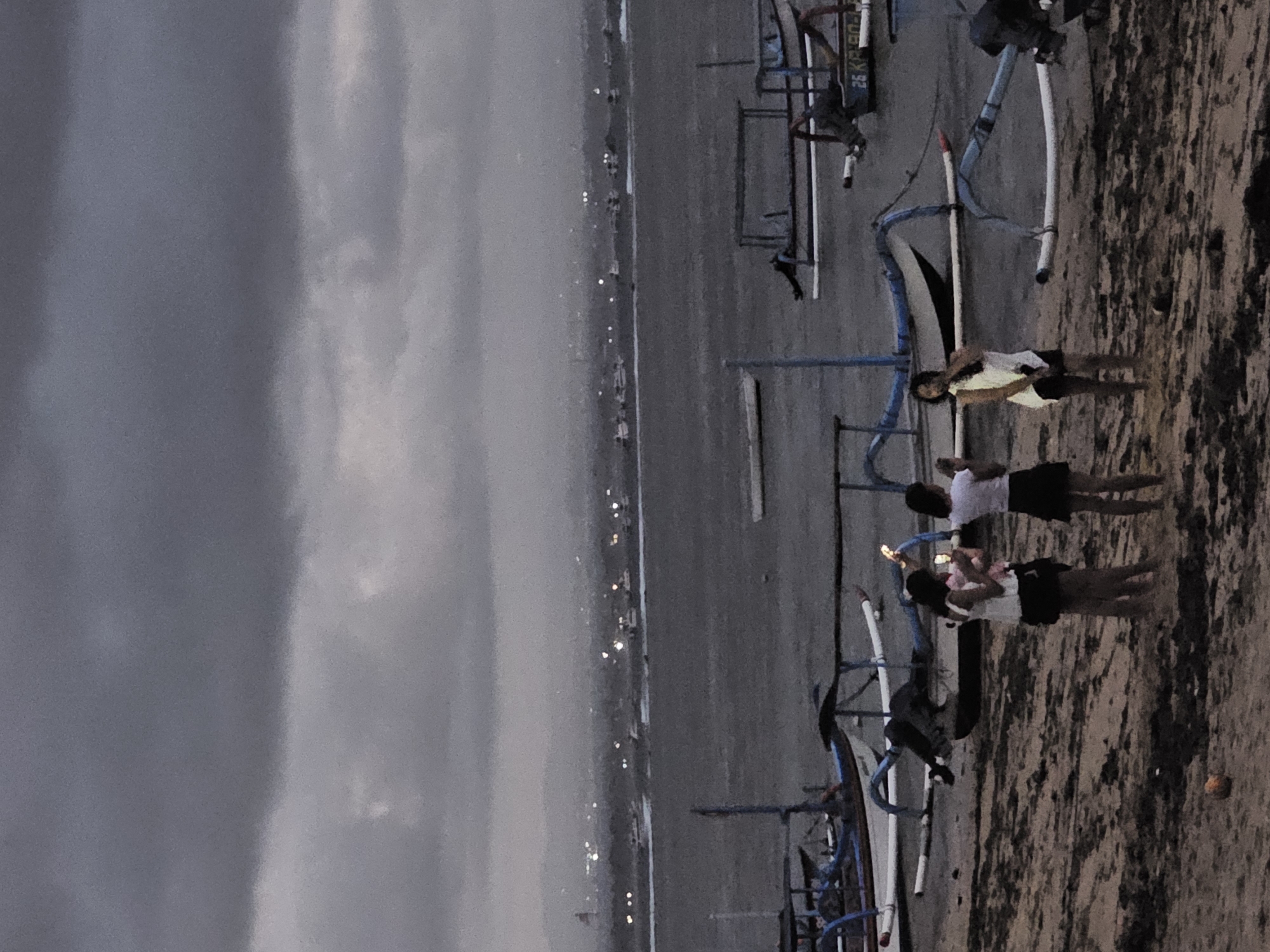 The image depicts a scene at Pantai Sanur, Denpasar, Indonesia, during what appears to be dusk or overcast conditions. In the foreground, three young women are standing on a sandy beach. The woman on the left is wearing a white top and a dark skirt, holding a phone and taking a picture. The middle woman, also in a white top and dark skirt, is holding a phone. The woman on the right is wearing a white top and dark shorts and is looking to her left. Behind the women, several traditional Balinese outrigger boats, known as jukung, are visible, some partially submerged in the water and others pulled up onto the shore. The water is a dark, choppy gray, indicating rougher conditions. In the background, a hazy cityscape with numerous lights is visible across the water, suggesting an urban area or developed coastline. The sky is heavily clouded and dark gray. The overall atmosphere is muted and somber due to the lighting and weather.