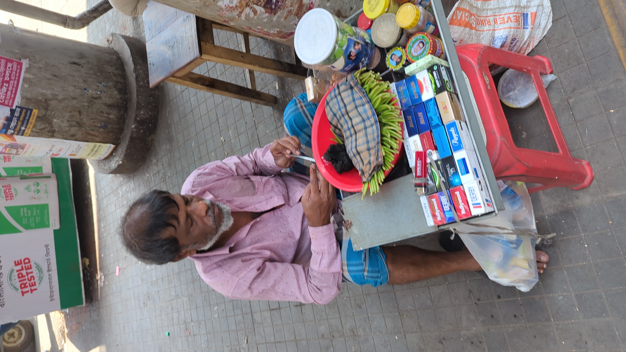 The image captures an outdoor scene in Dhaka, Bangladesh. An elderly man with a gray beard and balding head is seated on the ground. He wears a pink shirt, and a blue and white checkered lungi, and appears to be preparing a paan (betel leaf preparation). He holds a slim object, possibly a utensil, in his right hand.
Beside him is a makeshift stall constructed on what appears to be a gray metal tray. A red plastic basin sits atop it, containing green betel leaves and a checkered cloth. Various containers, jars, and packs of cigarettes are arranged on the tray. Brands such as Star, B&H, Regals, Camel, Derby, Hollywood, and Avon can be identified.
A red plastic stool is situated next to the tray, and a plastic bag is visible underneath. A wooden chair is positioned behind the stall, along with a large, cylindrical concrete post covered in posters, including one for a cement brand advertised as "Triple Tested".
The ground consists of gray paving stones. The scene suggests a typical street-side vendor.