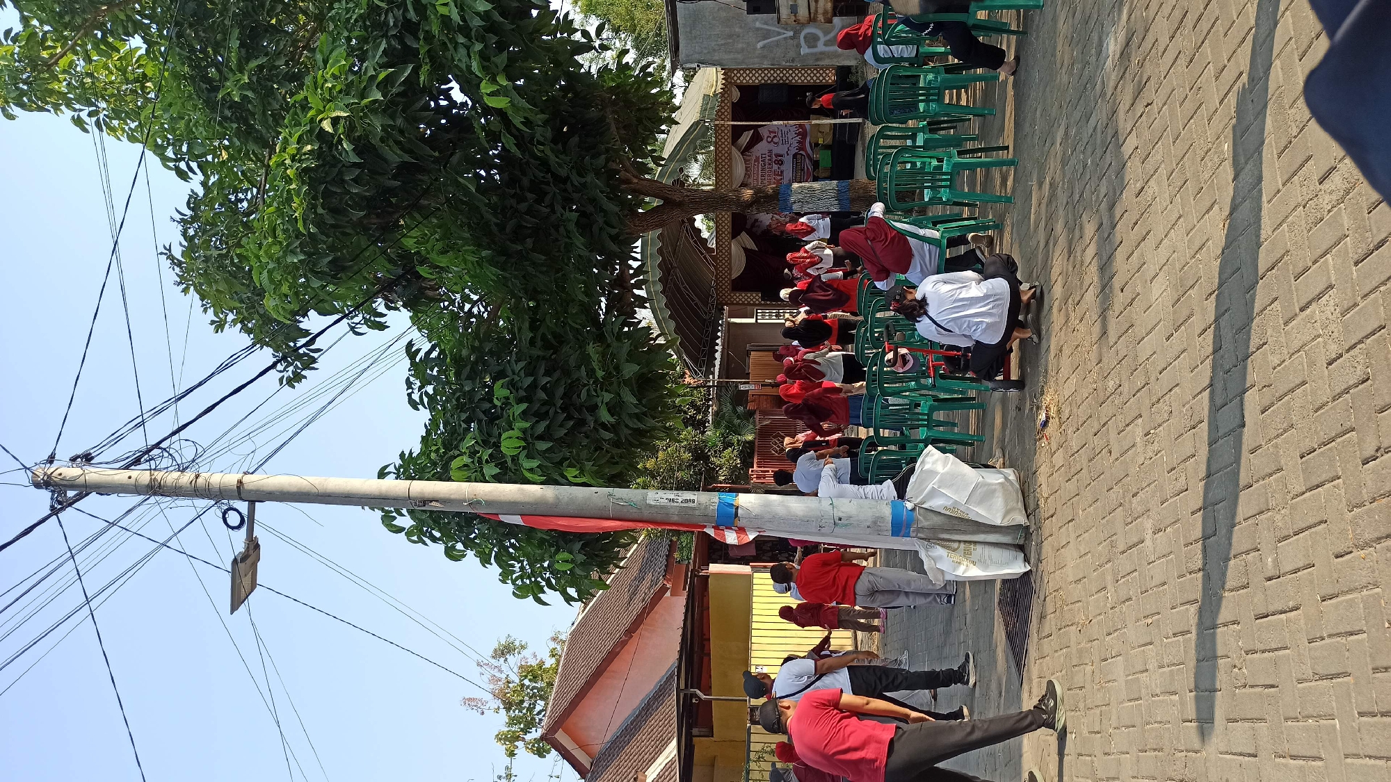 The image depicts an outdoor gathering of people in what appears to be a residential area in Galak, Indonesia. The scene is set during daylight hours, with clear skies suggesting a sunny day. In the foreground, a paved walkway with interlocking brick patterns is visible, with shadows cast by a tall utility pole and a large tree.
The utility pole, centrally located, is adorned with a red and white Indonesian flag and a collection of electrical wires. Near the base of the pole, two large white bags are placed on the ground.
A group of individuals, mostly dressed in red and white attire, are gathered in the mid-ground. Some are standing and appear to be engaged in an activity, possibly a morning exercise or a communal event, facing a makeshift stage or covered area. Others are seated on rows of green plastic chairs arranged in front of the stage. A few people are positioned in the foreground, observing the activity or resting.
The background reveals residential buildings with orange-tiled roofs and yellow-painted walls. A large, leafy tree with dense foliage provides shade over a portion of the gathering area. A stage or platform with a white canopy and decorative elements is set up, with banners displayed that are partially visible. The overall atmosphere suggests a community event or a local celebration.