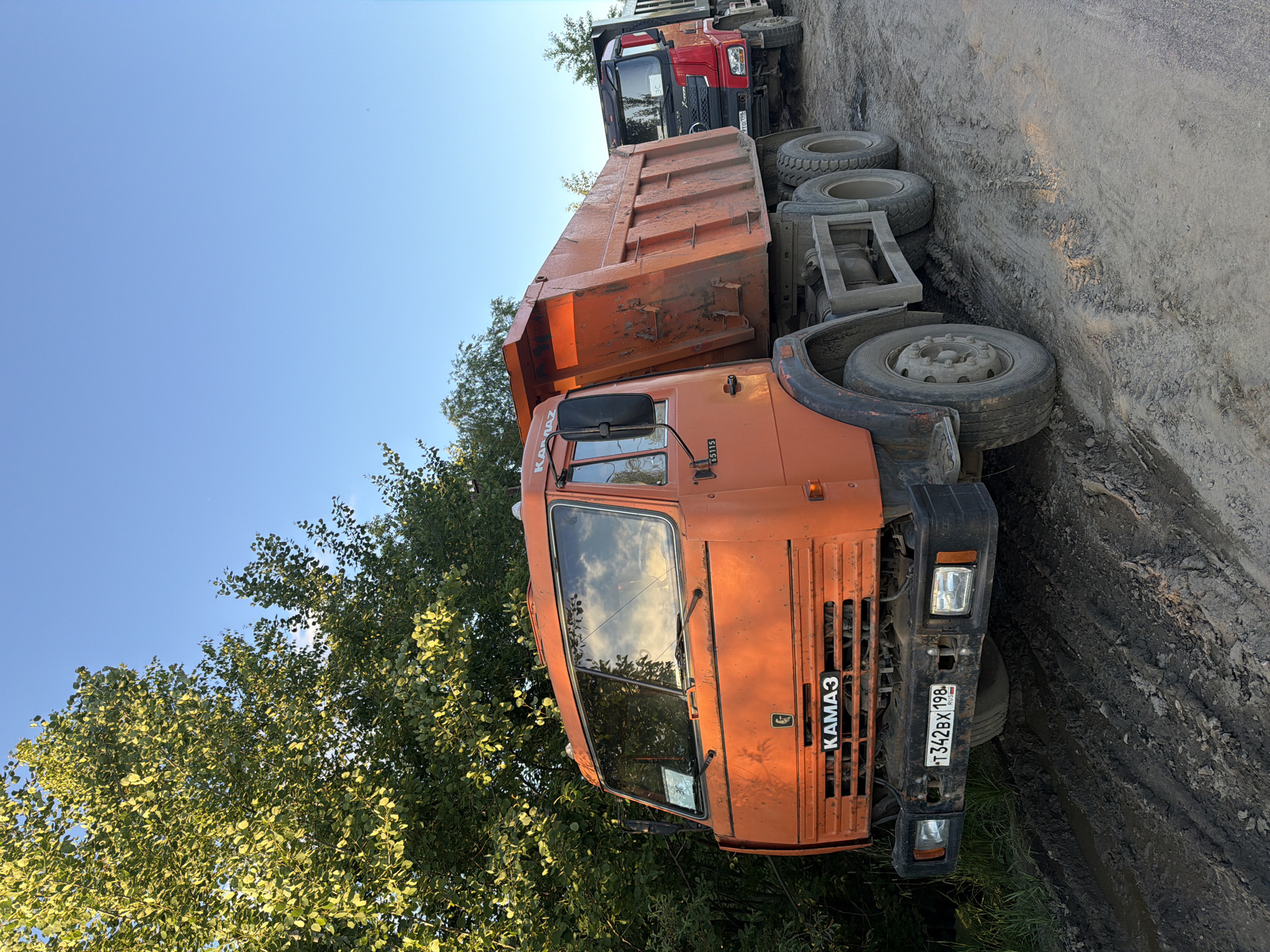 An orange dump truck is parked on a muddy dirt road next to a tree line. The truck has "KAMAZ" written on the front grill and a license plate that reads "T342BX 198 RUS". Another dump truck, red and black, is parked behind the orange truck. The sky is clear and blue.