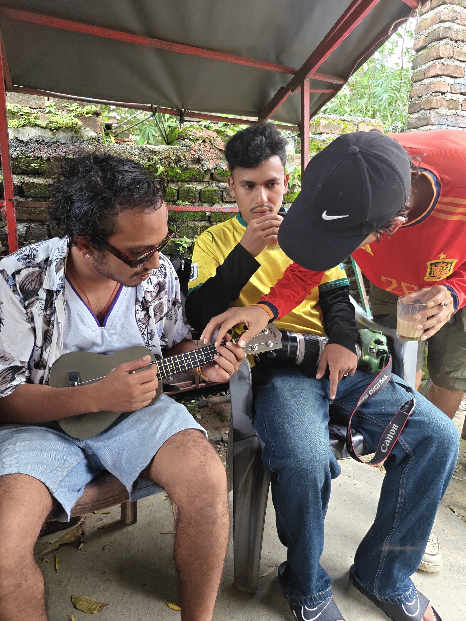 In Pokhara, Nepal, three young men are seated outdoors under a shaded structure. The man on the left, wearing a floral short-sleeved shirt over a white v-neck shirt and blue denim shorts, is playing a ukulele. He has dark, curly hair, sunglasses, and an earring. The man in the middle, wearing a yellow and green long-sleeved shirt with a black collar, looks down at the ukulele. The third man, on the right, is wearing a red long-sleeved shirt with a Spanish flag emblem on the sleeve and a black baseball cap with a Nike logo. He holds a clear plastic cup containing a light brown beverage. A professional camera with a long lens is positioned across the lap of the man in the middle, with a "Canon" branded strap visible. The background consists of a textured brick wall covered in moss and greenery, and foliage. The ground is concrete.