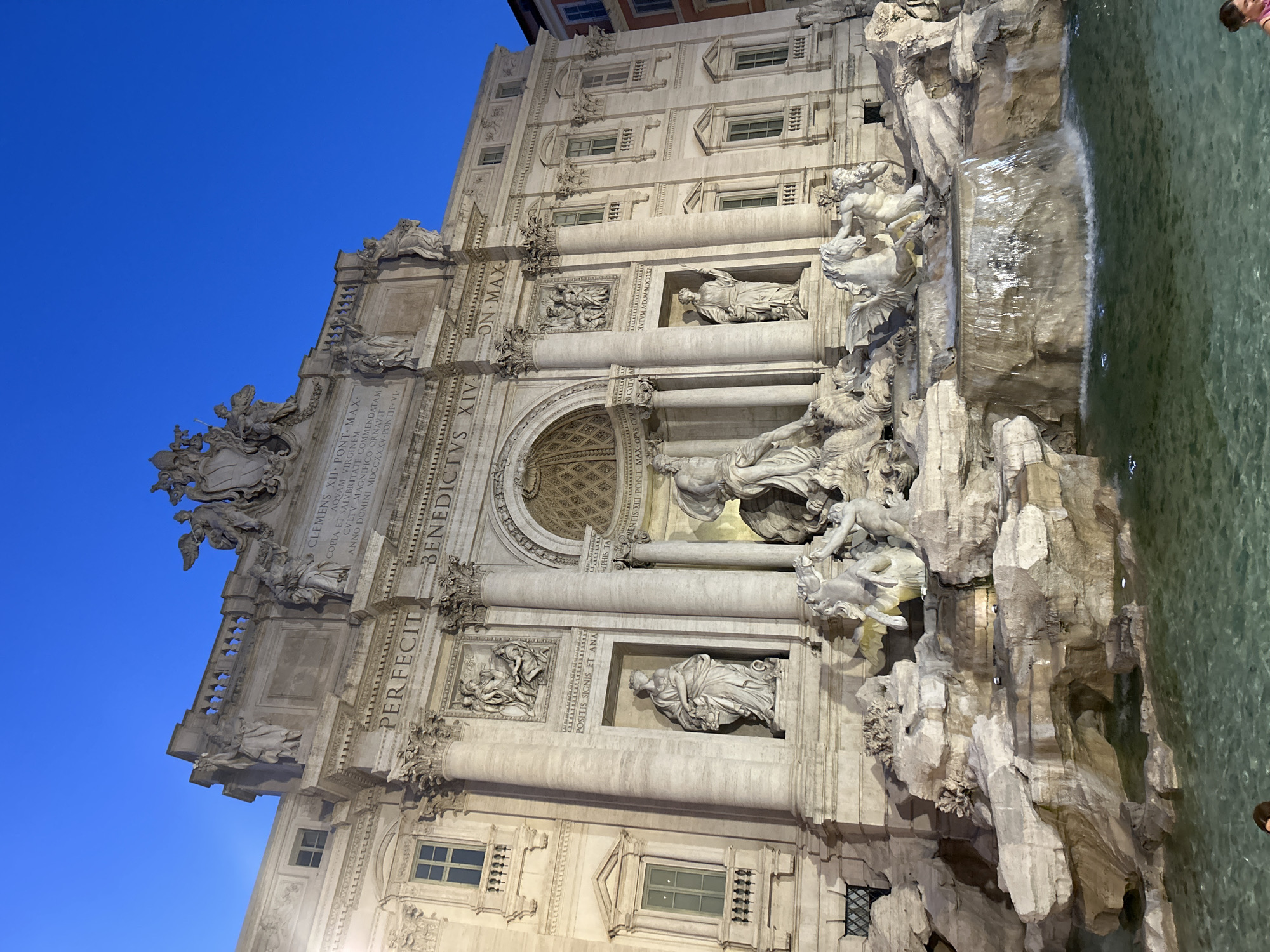 The image shows the Trevi Fountain, a famous landmark in Rome, Italy. The fountain is illuminated by artificial lights, highlighting its Baroque architecture and intricate sculptures. The water of the fountain is a clear green, with several cascades and pools. The sky above is a deep blue, indicating it is either dawn or dusk. The fountain is a large structure, with statues of gods and mythological figures, as well as horses and other animals. The scene is serene and majestic, with the sound of flowing water contributing to the ambiance.