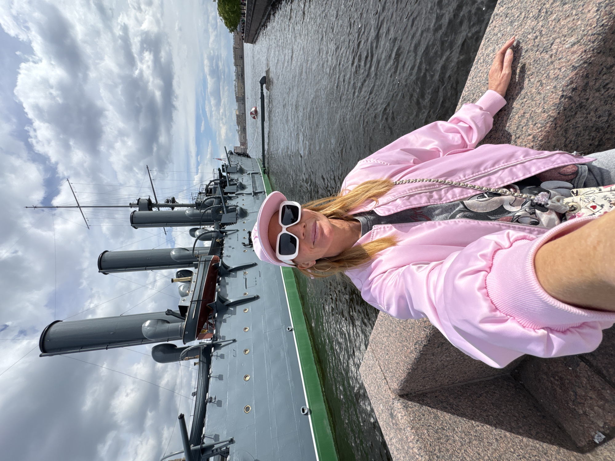 The image, oriented vertically and rotated 90 degrees clockwise from a typical horizontal view, features a fair-skinned woman posing outdoors on a bright, cloudy day. She has long blonde hair and wears a light pink bomber-style jacket, a matching pink baseball cap, and large white-framed sunglasses. Her right arm rests on a speckled reddish-brown granite embankment, and her expression is neutral to slightly smiling. A grey t-shirt with a graphic print is visible underneath her open jacket, along with a silver-colored chain or strap.
To her left and extending into the background is a large, grey warship, likely a historical cruiser such as the famous Cruiser Aurora, which places this scene in Saint Petersburg, Russia. The ship is docked on a body of water, presumably a river or canal, and features prominent grey funnels, masts, and visible gun emplacements. A distinct green stripe with a white line marks its waterline. Across the water, on the distant bank, a cityscape with buildings can be seen under a sky filled with scattered white and grey clouds. The water itself appears dark and slightly textured. No explicit text is visible in the image.