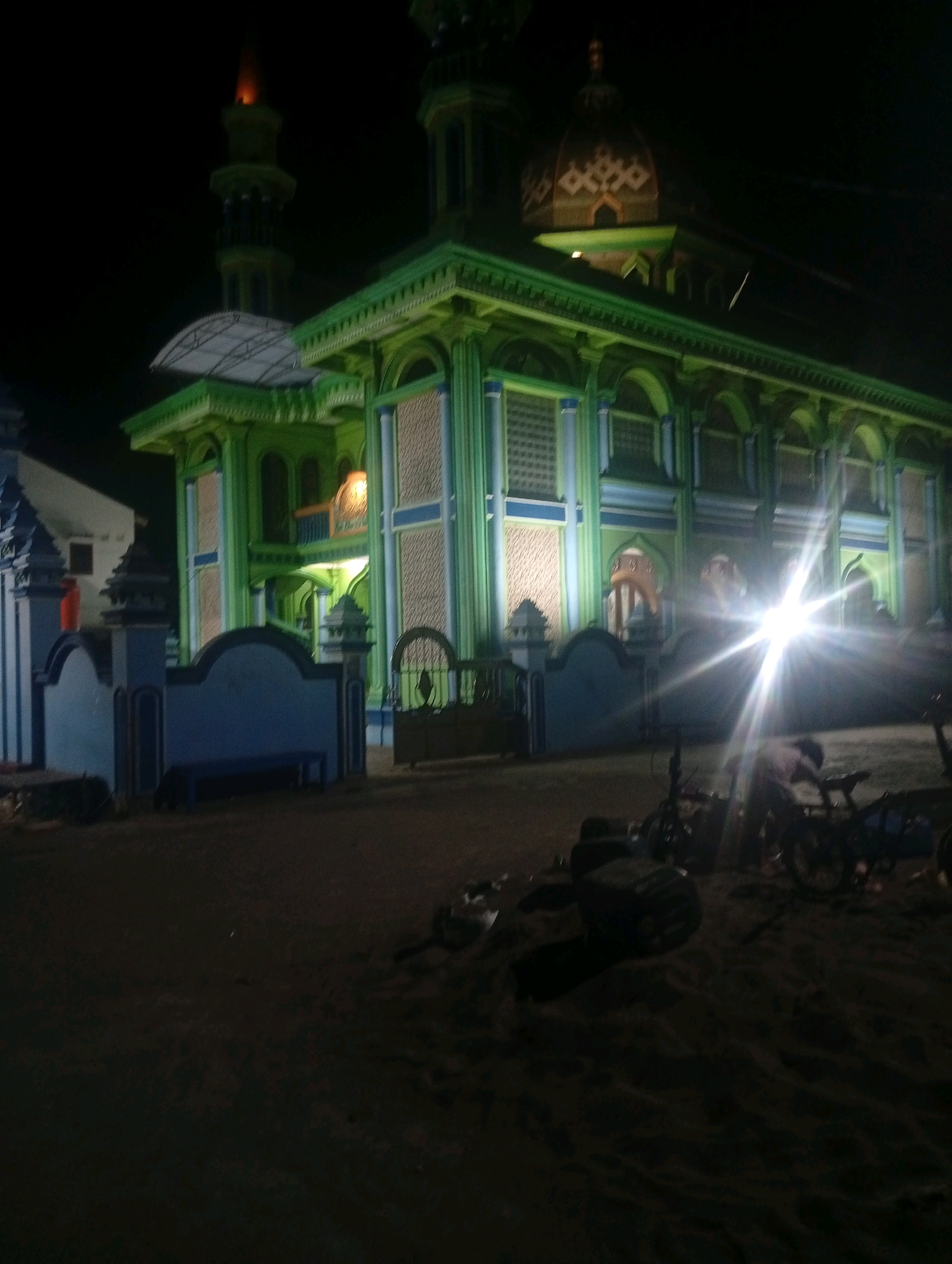 An exterior night shot of a brightly lit green mosque with blue accents is captured in Bajingjowo, Indonesia. The mosque features traditional Islamic architecture with a domed roof and a tall minaret. The surrounding area is dark and sandy, with some bicycles and debris scattered in the foreground. A few people are visible in the distance, one of whom appears to be working on a bicycle. The lighting is dramatic, with a strong lens flare from a bright light source in the center of the frame. The overall impression is one of a vibrant religious landmark set against a simple, somewhat unkempt, natural environment.