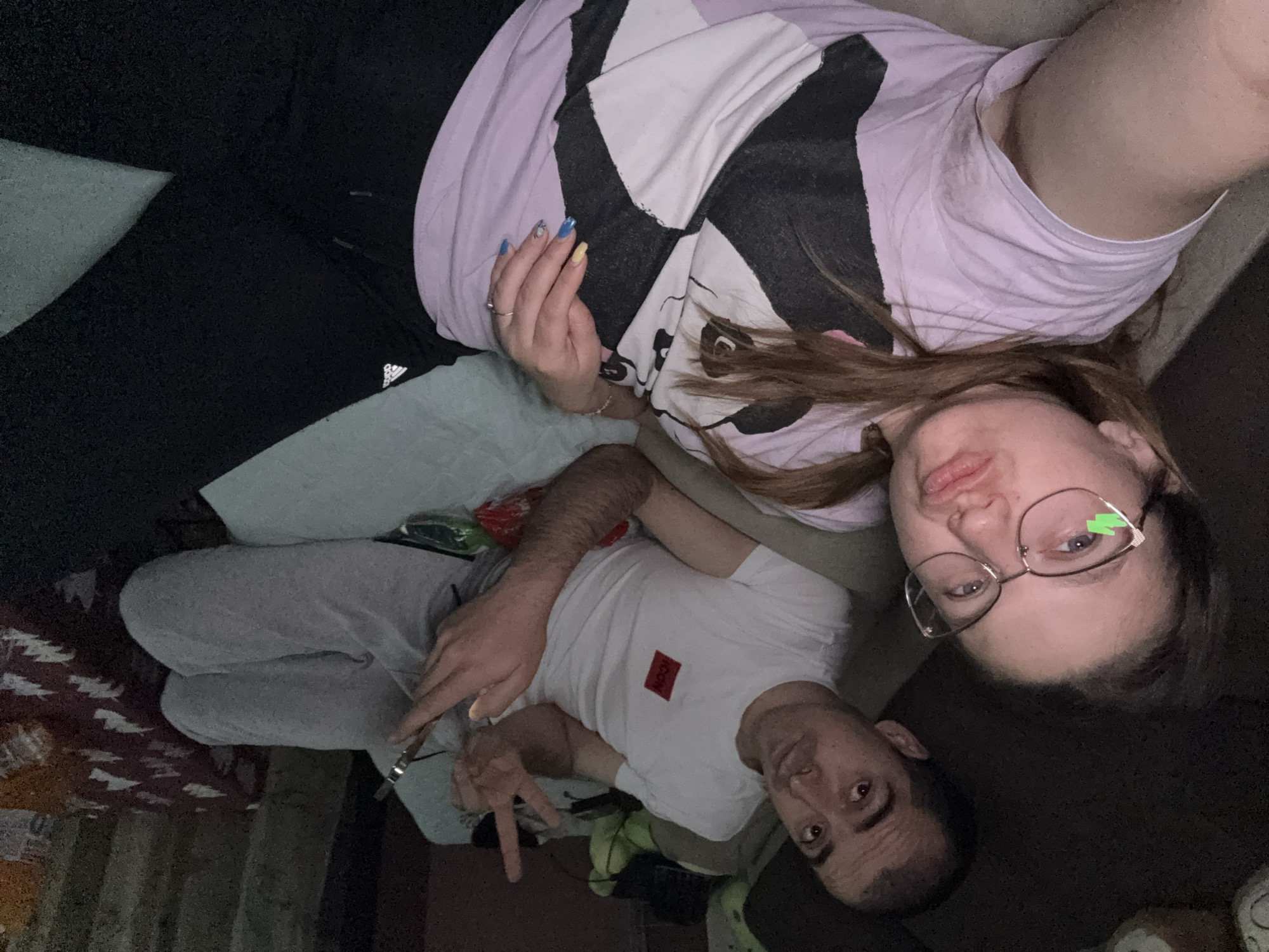 Two individuals are visible in an indoor setting. A woman wearing glasses and a t-shirt with a black-and-white design and a lavender hue is positioned at the top of the frame. A man wearing a white t-shirt with a red square logo is below her. The setting appears to be a couch or bed. The location is unknown and no evidence exists in the image that can confirm that this image was taken in Sofia, Bulgaria.