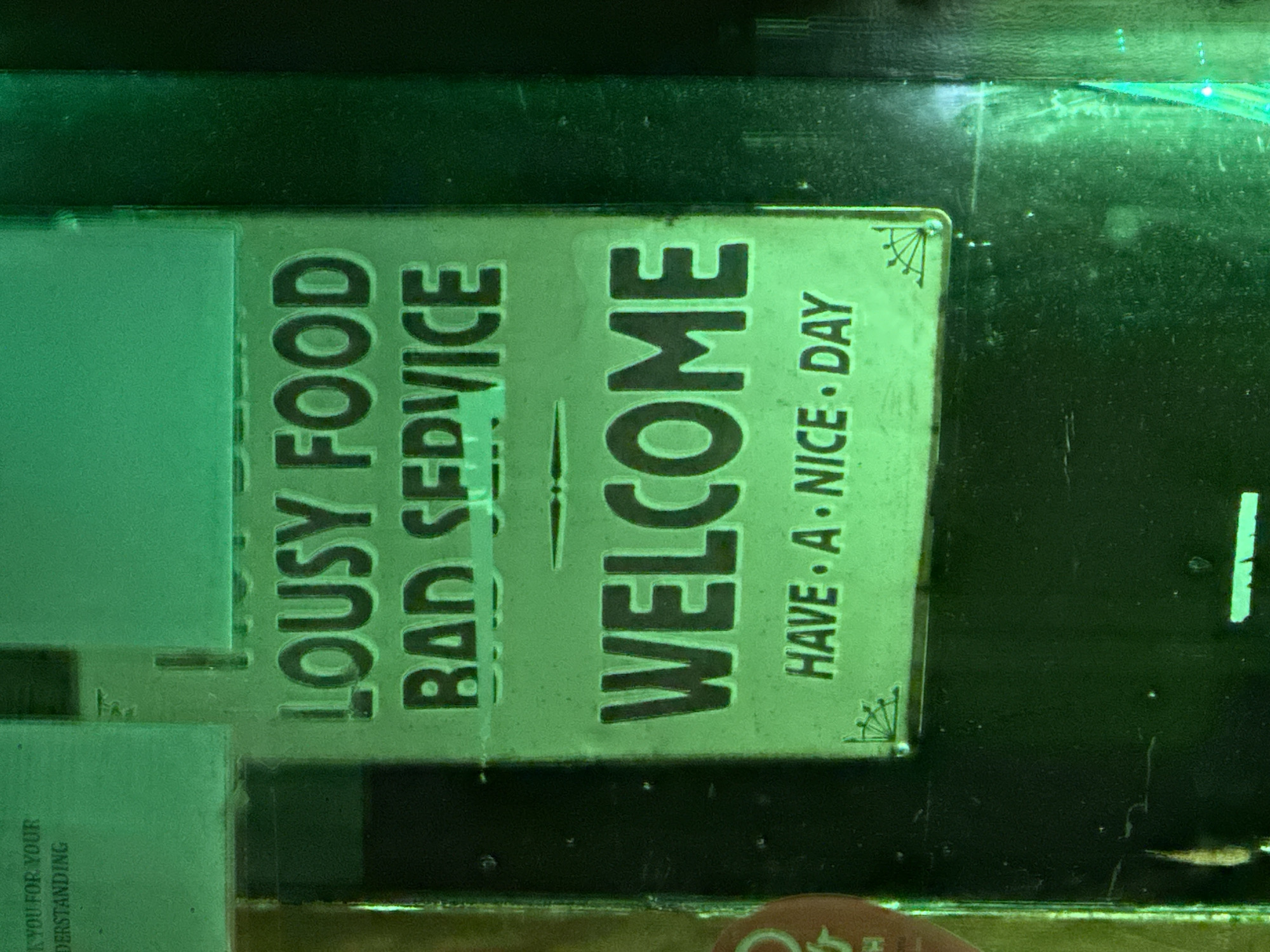 The image shows a humorous metal sign, likely displayed inside a business. It reads "LOUSY FOOD, BAD SERVICE, WELCOME, HAVE A NICE DAY." The sign appears to be attached to a surface, perhaps a cabinet or shelf. The image has a green tint, possibly due to the lighting. I cannot determine the location in Amsterdam, Netherlands from this single image.