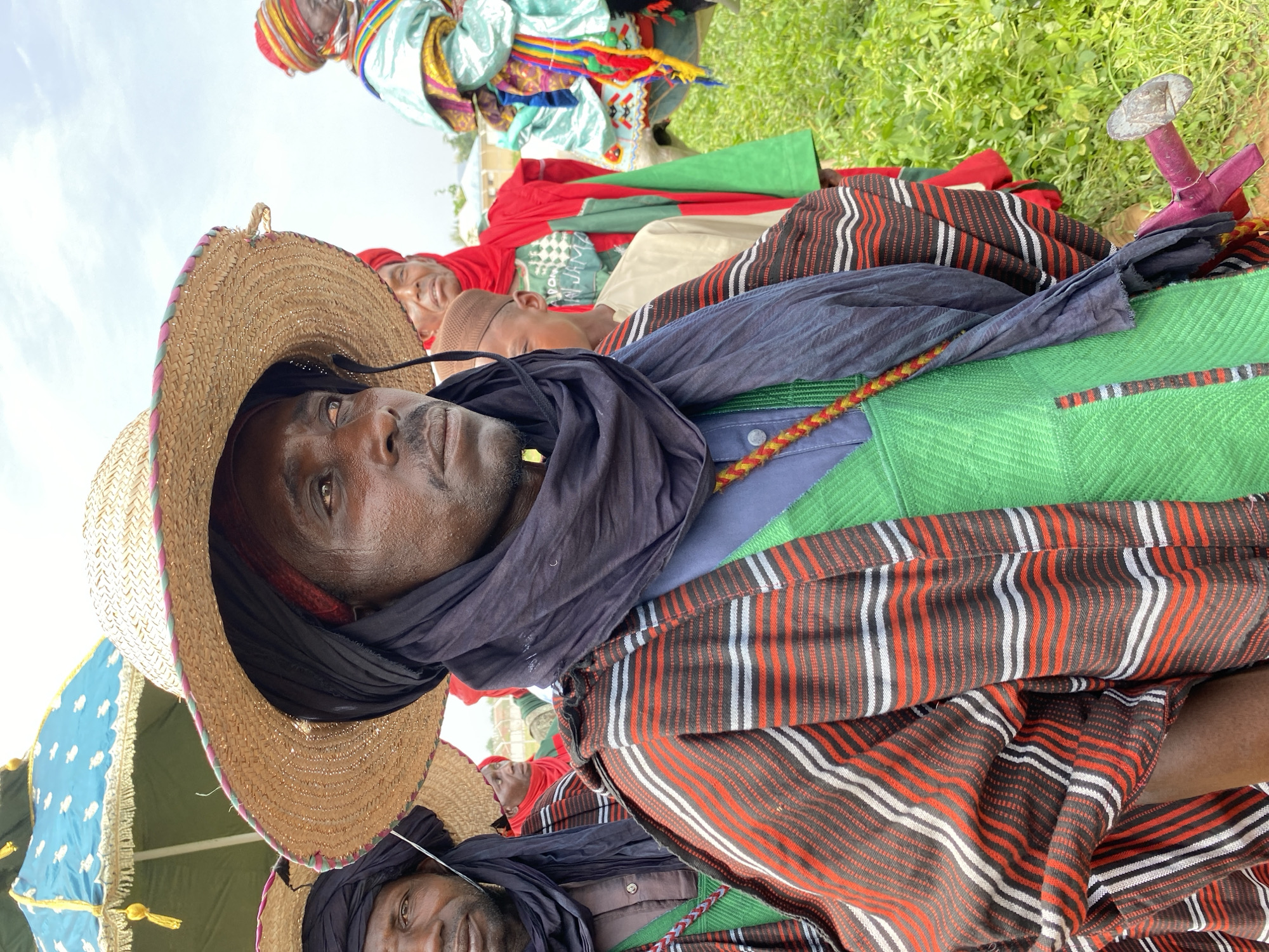 A close-up, low-angle, and slightly distorted portrait shows several individuals resting on the ground. A man in the foreground wears a straw hat with a colored brim and a dark head wrap or scarf around his neck. He is dressed in a light blue shirt, a green jacket, and a horizontally striped red, white, and black patterned textile. Behind him, others are lying down, some in red fabric, and one wearing a multicolored head covering. Green grass is visible in the background. A small metal object sits on the ground next to the subjects. The photo appears to have been taken in a field setting. The scene is likely a resting period during an event. Kafanchan A, Nigeria, is only provided as the location.