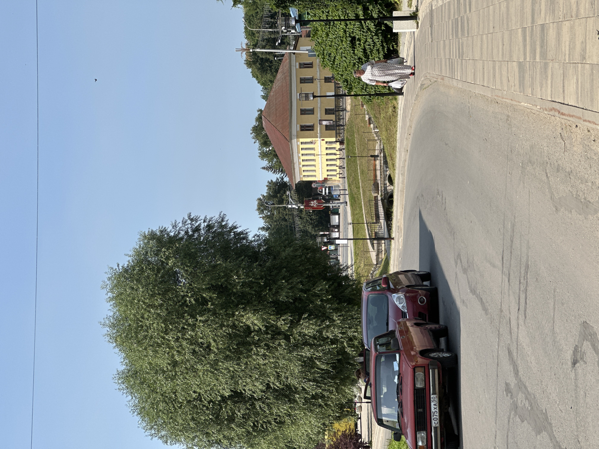 A sunny day scene captures a curved street in the city of Terpilovo, Russia. Two red cars are parked along the road, with one a classic sedan and the other a more modern hatchback. A large, leafy tree dominates the left side of the frame, casting dappled shadows. In the background, a yellow, two-story building with a red roof stands against a clear blue sky. A woman in a long, patterned dress walks along the tiled sidewalk, carrying two plastic bags. The street appears to be well-maintained, with a pedestrian crossing and lampposts lining the sidewalk. The overall impression is one of a peaceful, everyday scene in a Russian provincial town.