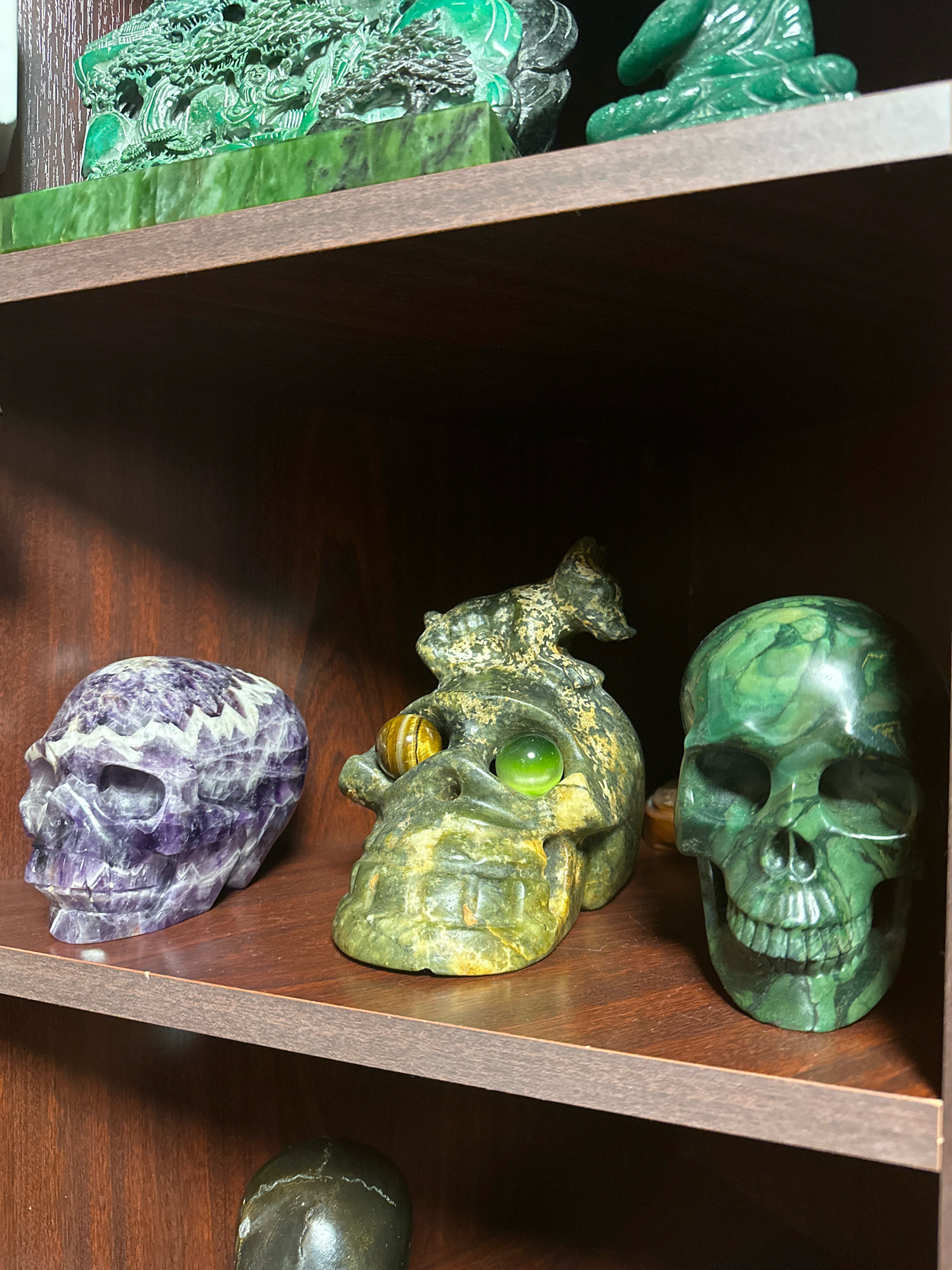 The image shows a close-up view of a wooden shelf containing several carved stone objects. On the top shelf, a large, intricately carved green jade sculpture depicting a landscape with trees and figures is visible. To the right of this, a smaller green jade carving of a seated Buddha is present.
The middle shelf holds three carved stone skulls. On the left is a purple and white banded amethyst skull. In the center is a greenish-yellow stone skull with a creature perched on top, featuring a tiger's eye stone and a green spherical stone embedded in its forehead. To the right is a vibrant green stone skull with pronounced features and carved teeth.
The bottom shelf is partially visible and contains a dark, possibly stone, spherical object at the lower left corner. The background is a dark wooden cabinet. The image appears to be taken indoors in a dimly lit environment. There is no information to place this image in Murmansk, Russia.