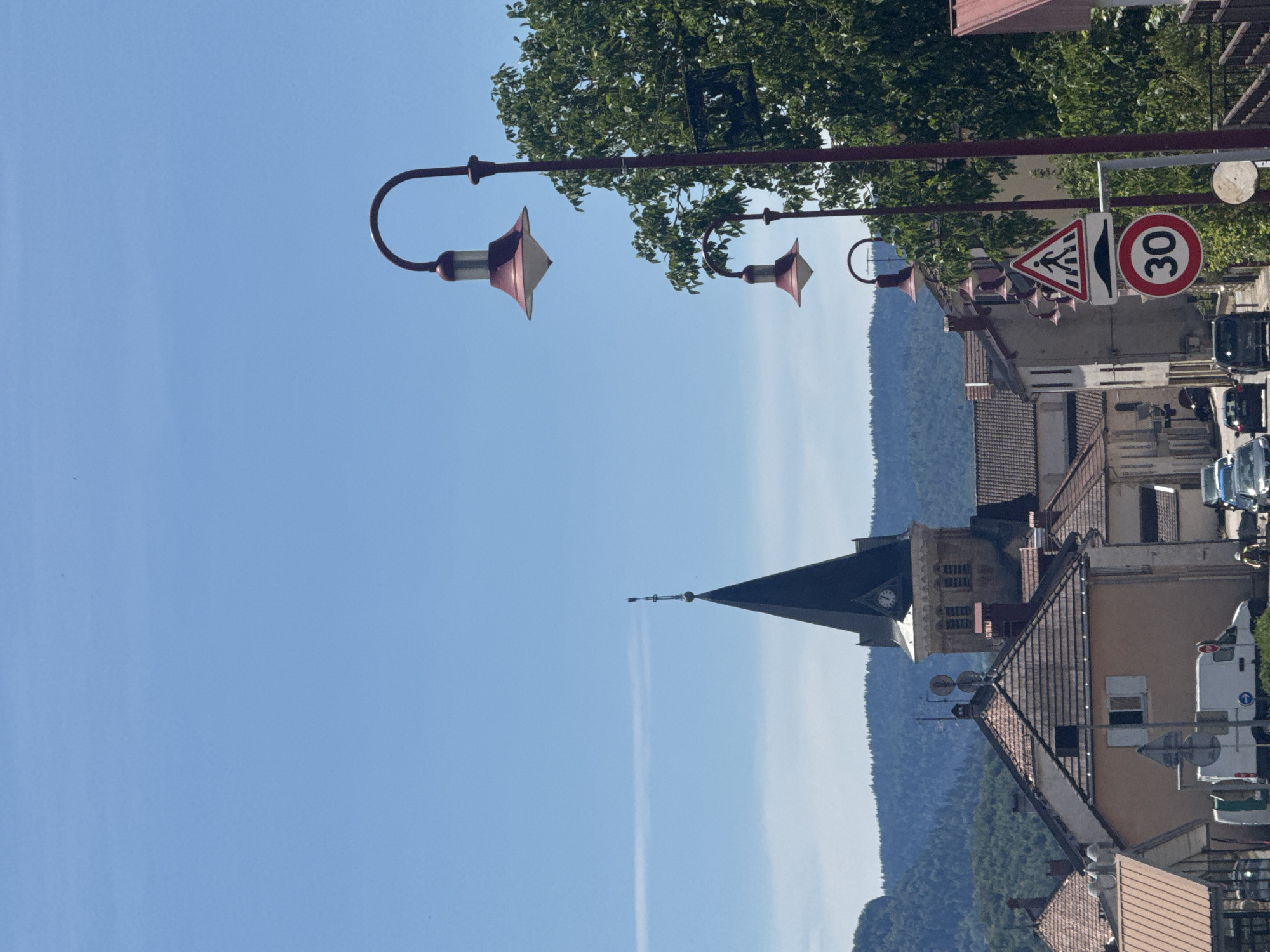 The image depicts a street scene in the town of Coteaux du Lizon, France, under a clear blue sky. In the foreground, a tall, reddish-brown lamppost with three illuminated lamps is positioned to the right of the frame. Behind it, a large, leafy green tree dominates the right side of the image. To the left and center, a cluster of buildings with terracotta roofs are visible. A prominent church spire, dark in color, rises above the rooftops in the middle ground, featuring a clock face. In the distance, rolling green hills form a backdrop.
Several vehicles are present on the street, including a white van with a blue arrow decal on its rear door, and other cars further down the road. Traffic signs are also visible: a pedestrian crossing sign with a speed bump symbol below it, and a circular "30" speed limit sign. The architecture suggests a European village setting. No people are clearly identifiable in the scene, though a figure wearing a yellow vest is partially visible near the vehicles. The lighting indicates it is daytime, likely late morning or early afternoon.
