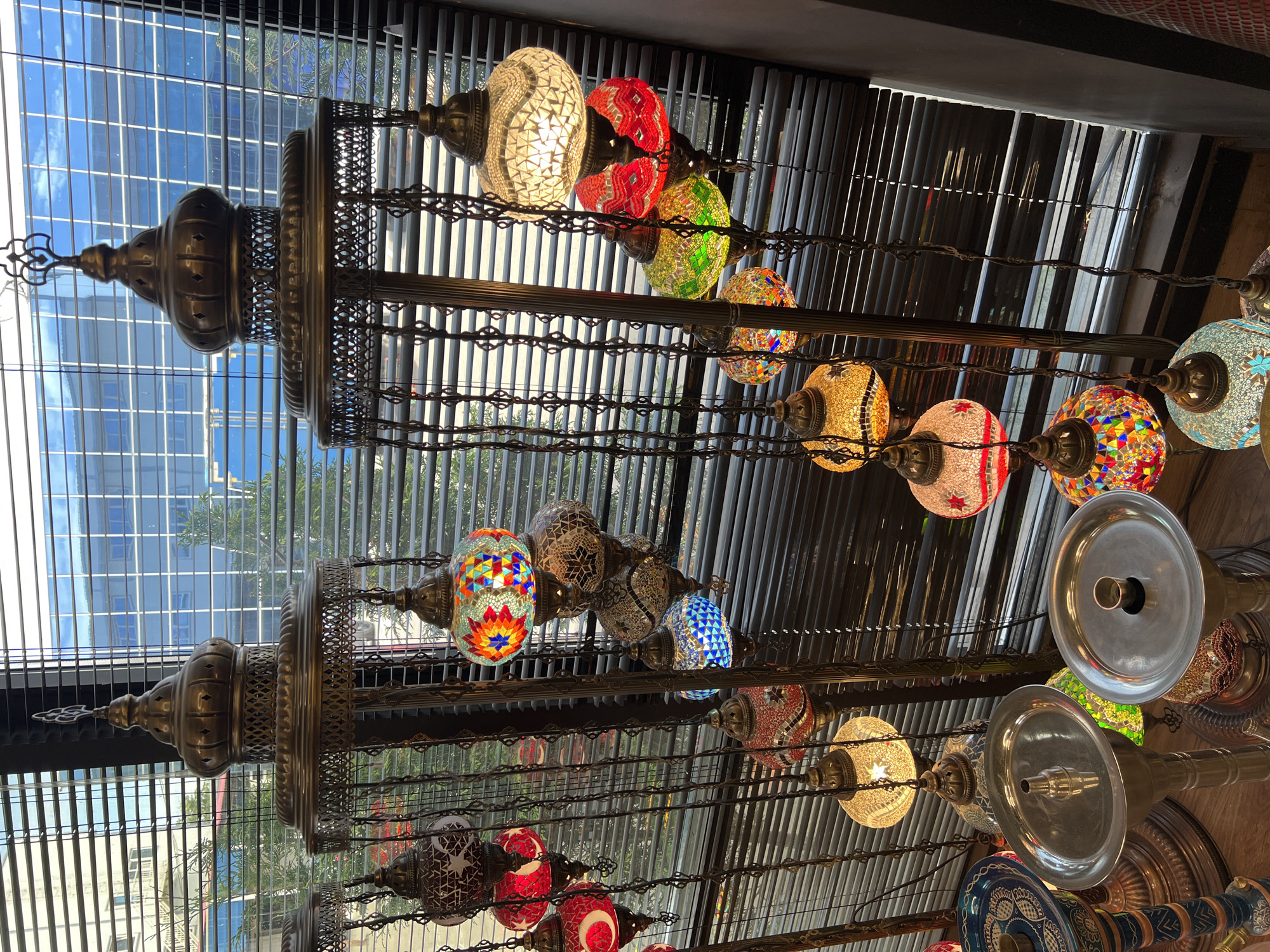 The image displays an indoor scene featuring numerous ornate, colorful mosaic lamps of varying sizes, arranged vertically along two large, dark metal decorative fixtures or chandeliers. These lamps, reminiscent of Turkish or Moroccan designs, are intricately crafted with small, brightly colored pieces of glass, some forming patterns like stars or geometric shapes. Many of the lamps are illuminated, casting a warm, inviting glow.
Behind and above the hanging lamps, a large window is visible, covered partially by horizontal blinds. Through the window, a clear blue sky with some white clouds, parts of modern buildings with grid-like windows, and glimpses of green trees are discernible, indicating a daytime setting. The view suggests an urban environment.
In the bottom right corner of the image, several metallic, dish-shaped objects, possibly parts of hookahs or other decorative items, are also visible, contributing to the exotic ambiance. The overall impression is that of a shop, market, or restaurant specializing in or decorated with Middle Eastern-inspired decor. No people or visible text are present in the image.