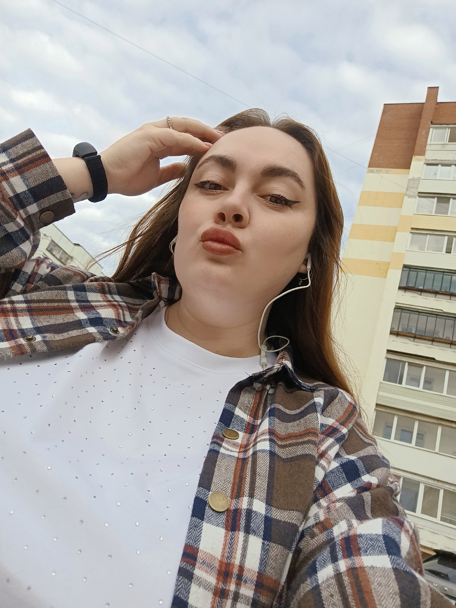 A woman is standing outside in Chaikovsky, Russia, taking a selfie. She is wearing a white t-shirt covered with small embellishments, a brown and blue plaid shirt jacket, and a black watch on her left wrist. She has brown hair, dark eyeliner, and is pursing her lips in a pout. In the background, a tall apartment building with yellow and white stripes, brick red trim, and several windows is visible. The sky is cloudy.