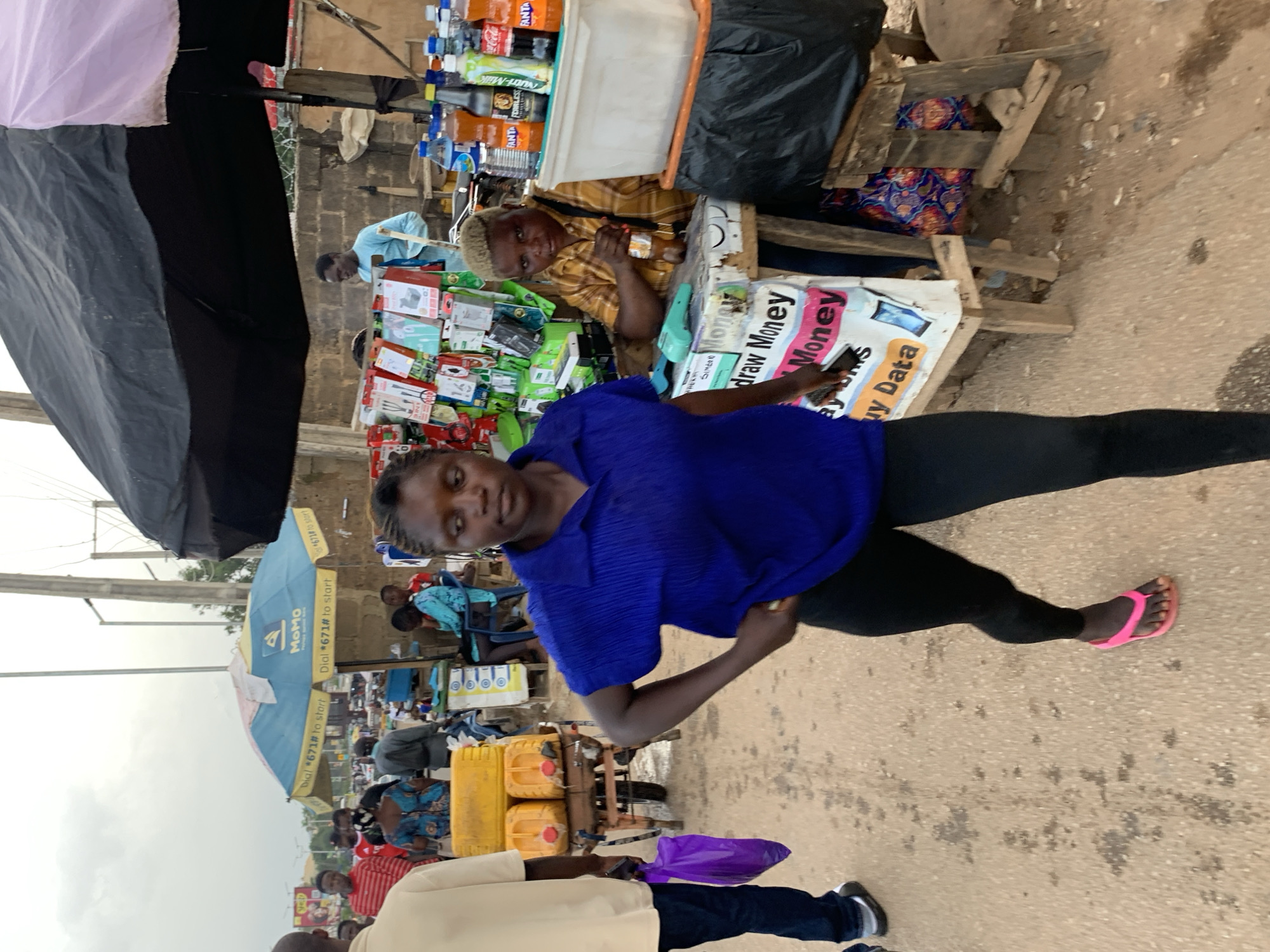 The image depicts an outdoor market scene in Jos, Nigeria, with a focus on a woman walking towards the camera. She is wearing a bright blue collared shirt and black leggings, with pink flip-flops on her feet. She has her left hand on her hip, and appears to be looking forward. To her right, a person with short, blonde hair is seated behind a stall, holding a bottle and looking towards the camera. The stall is covered by a large black and pink umbrella and displays various electronic accessories and bottled drinks. Behind the stall, a cart with yellow jerrycans is visible, and other people are seen in the background, some under a blue and yellow umbrella. The ground is dusty and appears to be a dirt road, with some scattered debris. The overall atmosphere suggests a busy, informal marketplace.