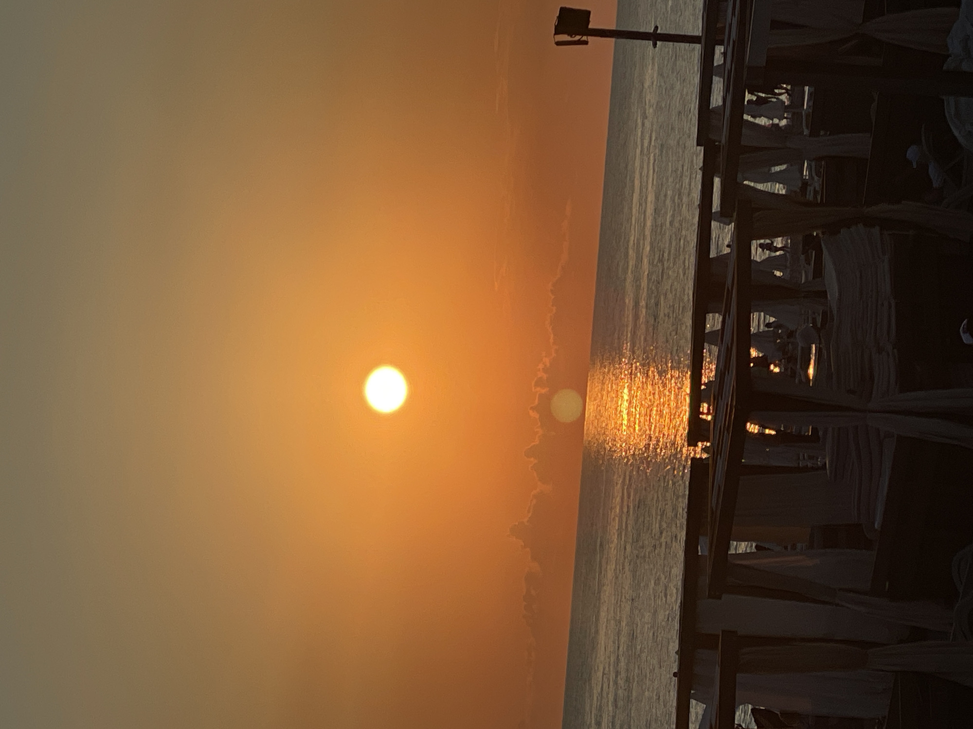 The image depicts a sunset over a body of water, likely the sea, with a pier or boardwalk in the foreground. The sun, a bright orange orb, is low in the sky, casting a warm glow and creating a shimmering reflection on the water's surface. Wispy clouds are visible, catching the last rays of sunlight. The pier is constructed of dark wood and features several white cabana-like structures, suggesting a resort or recreational area. The lighting is soft and atmospheric, indicative of either sunrise or sunset. The overall mood is serene and tranquil. The image appears to be taken in Sochi, Russia, based on the provided context. No visible text is present in the image.