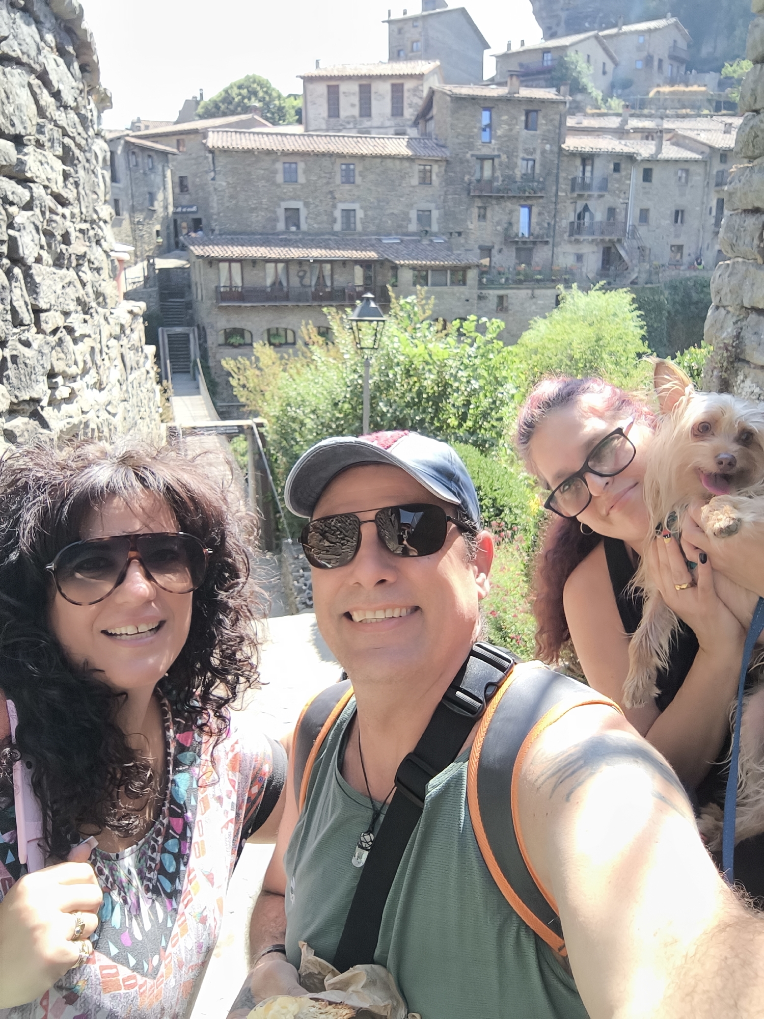 This is a selfie taken by a group of three people, two women and one man, with a small dog, in a town with stone buildings, possibly Rupit i Pruit, Spain. The man, in the foreground and slightly off-center to the right, is wearing sunglasses, a cap, and a green t-shirt with a backpack. The woman to his left is wearing large sunglasses and a colorful patterned top. Behind them, a second woman, on the right, is holding the small, light-colored dog, and is wearing glasses and a black top. The background shows a picturesque village scene with stone houses, tiled roofs, and lush greenery, suggesting a sunny day. The people appear to be posing for the camera and smiling.