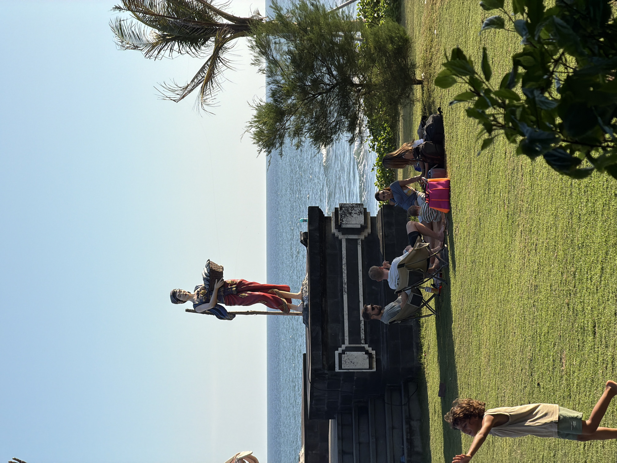 A statue of a woman holding a basket stands on a raised platform by the ocean in Mengwi, Indonesia. The statue depicts the woman in traditional attire, with a red sarong and blue top. She holds a woven basket filled with what appear to be fish. Below the platform, a group of people are relaxing on the grass. Two men sit in camping chairs, facing away from the camera. Further to the right, several other individuals, including a child, are seated on the ground, seemingly enjoying a picnic. The ocean stretches out behind them, with gentle waves lapping at the shore. A palm tree and other foliage frame the scene, suggesting a tropical coastal environment. A young child is in the foreground, running and waving their arms.