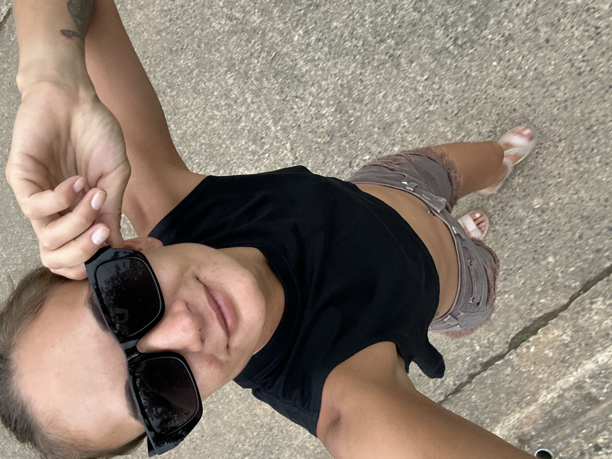 A young woman is standing on a paved surface, possibly a sidewalk or street, outdoors. She is wearing a black crop top, denim shorts with a frayed hem, and translucent sandals. She is holding up a pair of black sunglasses with her right hand, which is resting on her forehead. Her left arm is extended downwards, and her hand is resting on her hip. She is looking towards the camera and appears to be smiling faintly. The background is blurred, but it appears to be an outdoor environment with a grayish, textured ground. No specific landmarks or elements of Kathu, Thailand, are identifiable in the image.