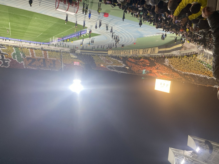 This image depicts a vibrant night scene inside the Complexe Sportif de Fès stadium in Fez, Morocco. Thousands of spectators fill the multi-tiered stands under bright artificial lighting.
A large-scale tifo display dominates the far side of the stadium, formed by the crowd holding colored cards or flags. This display prominently spells out "FEZ" in large yellow characters against a darker background, flanked by other intricate patterns in yellow, black, red, and white. Below this tifo, a long banner stretches across a section of the stand, displaying the text "WE ARE YOUR HUCKLEBERRY."
A bright scoreboard on the upper left of the far stand shows "0:00." The green football pitch with its white markings and goalposts is clearly visible, surrounded by a blue running track. Scattered individuals, identified as security personnel or event staff by their uniforms (some wearing reflective vests), are positioned along the track and near the pitch.
In the foreground, a dense crowd of spectators, many dressed in yellow and black attire, occupy the lower stadium seats. Numerous individuals in this section are holding up smartphones, actively recording or photographing the event. An overhead structure, possibly a part of the stadium's lighting rig or roof, is visible in the upper left corner, while a bright, glowing stadium floodlight dominates the upper right, casting a strong glare. A metal railing is partially visible in the immediate foreground, indicating the elevated perspective of the shot.