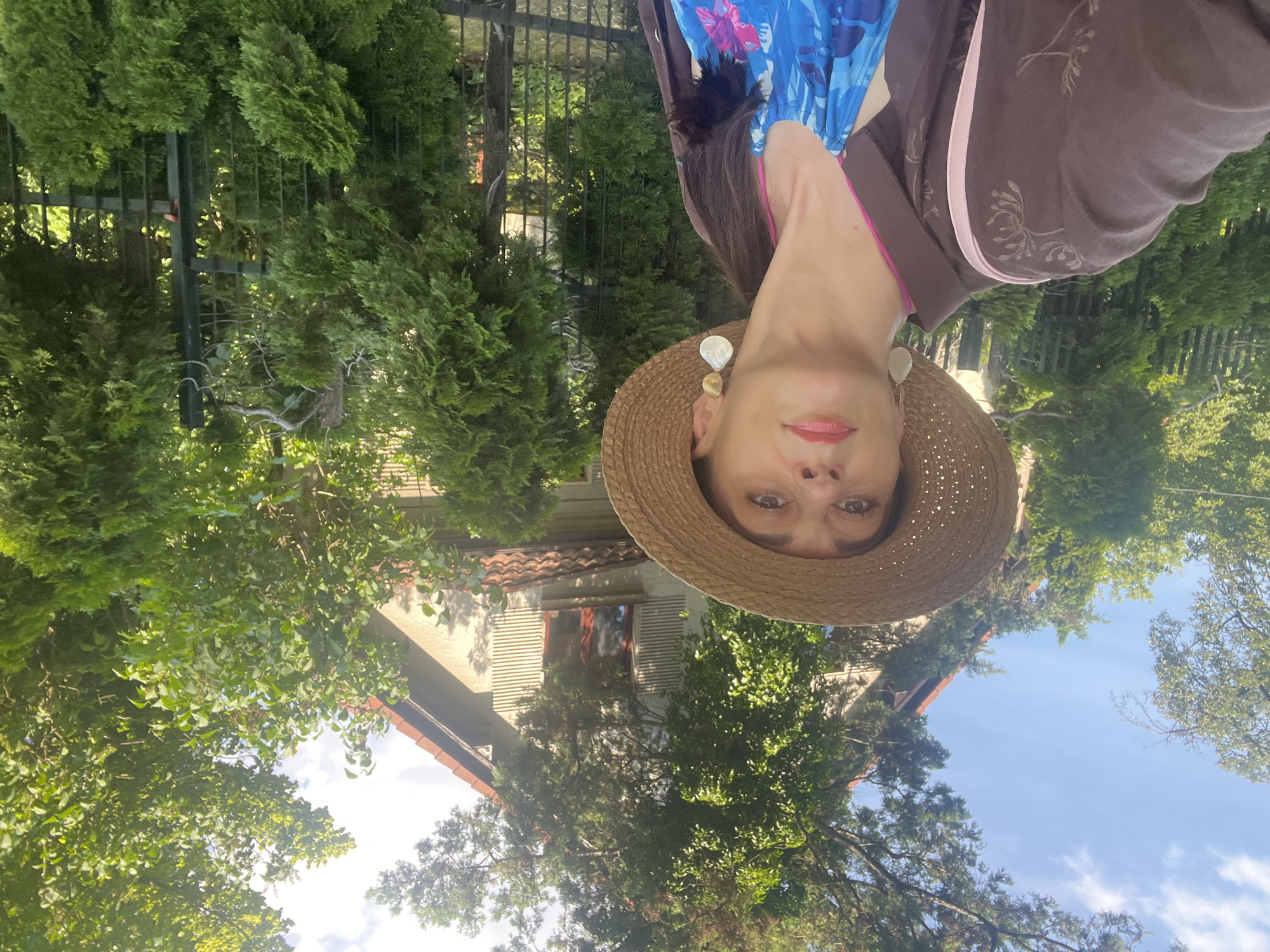 A medium shot shows a woman with brown hair and brown eyes wearing a straw hat, large teardrop earrings, a brown jacket, and a blue floral dress. She is taking a selfie outdoors in front of a house with a red-tiled roof and trees. The woman is looking directly at the camera. The house has a light-colored facade with some windows visible. There is a black metal fence in the foreground and lush green foliage surrounding the scene. The sky is blue with some white clouds. The overall lighting suggests it is daytime.