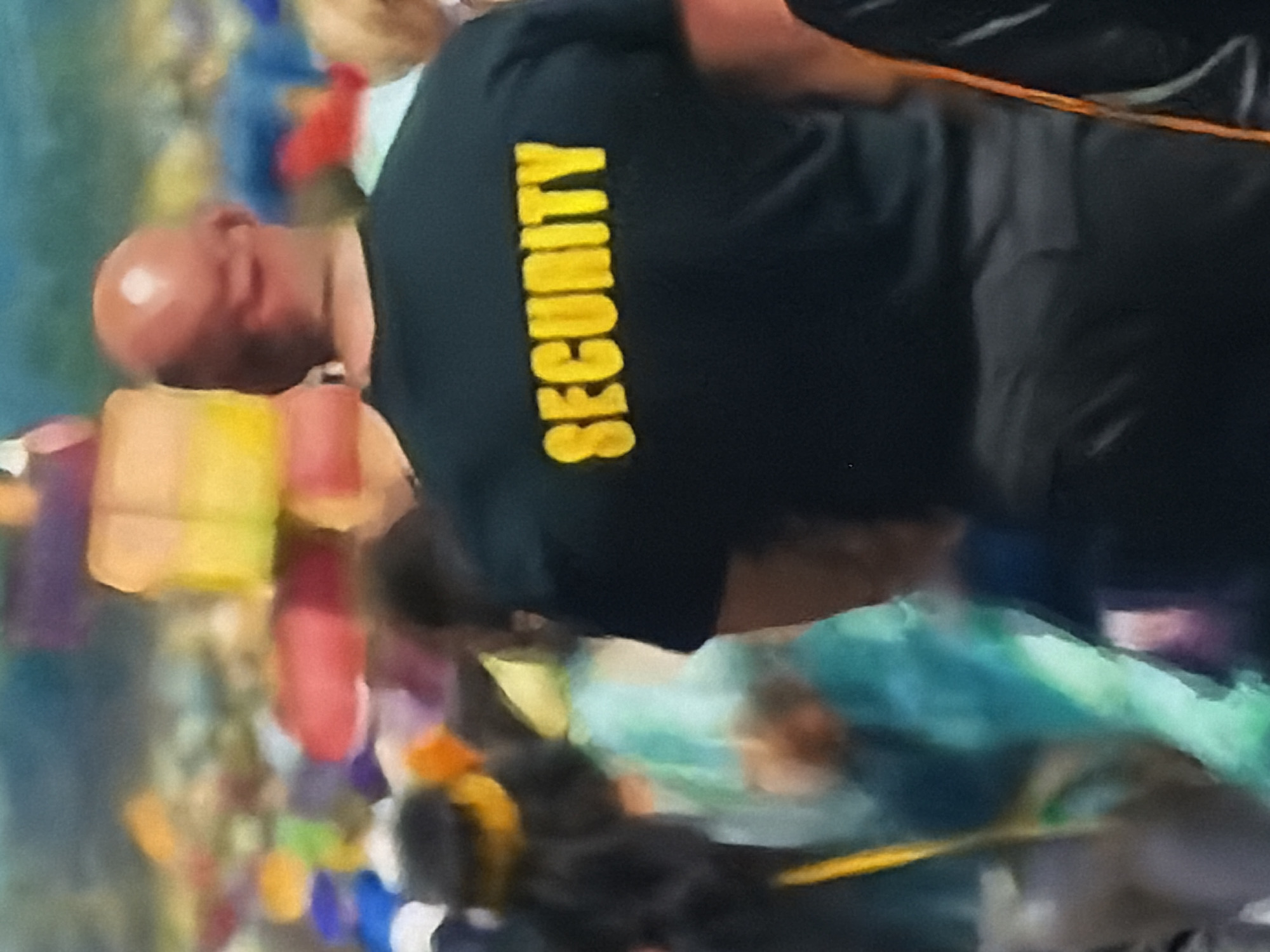 The image depicts a security guard from behind, standing in what appears to be a busy, colorful indoor public venue in Sofiivska Borshchahivka, Ukraine. The primary subject is a bald male wearing a black short-sleeved t-shirt. The word "SECURITY" is prominently printed vertically in bright yellow capital letters on the back of his shirt.
The background is largely out of focus but reveals a vibrant and active scene. Numerous blurry shapes and colors, including shades of yellow, red, blue, and green, suggest the presence of other people—possibly a crowd or gathering, some of whom may be children given the colorful elements—and various decorations or displays. The overall setting implies a lively event or location such as an entertainment center, a children's play area, or a public exhibition. The security guard's presence indicates supervision of the area. The lighting appears to be artificial, consistent with an indoor environment. No specific weather conditions are discernible, and individual emotions are not visible due to the angle and blurriness of the other subjects.
