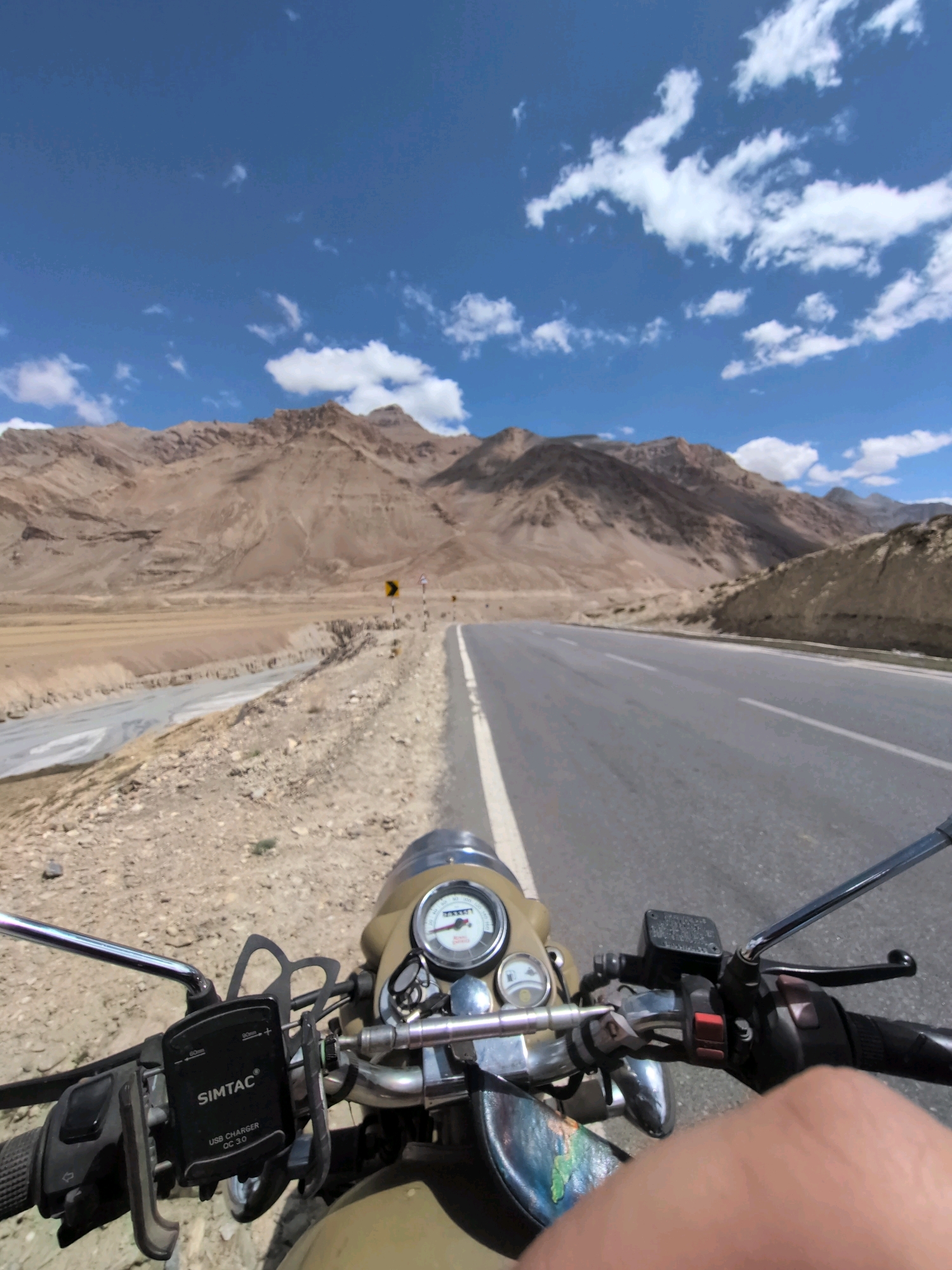 An extreme point of view, first-person perspective shot captures a motorcycle rider's view while traveling on a paved road in a mountainous, arid region. The rider's hand is visible on the right side of the frame, suggesting active control of the vehicle. The motorcycle's handlebars, mirrors, and dashboard with a speedometer are prominently displayed in the foreground. A SIMTAC USB charger is mounted on the handlebars. The road stretches ahead, bordered by rocky terrain and a dry riverbed. In the distance, rugged, brown mountains rise against a bright blue sky dotted with white clouds. The scene suggests a remote, possibly high-altitude location, consistent with regions like Dhar Sajam, India, known for its mountainous landscapes and scenic routes.