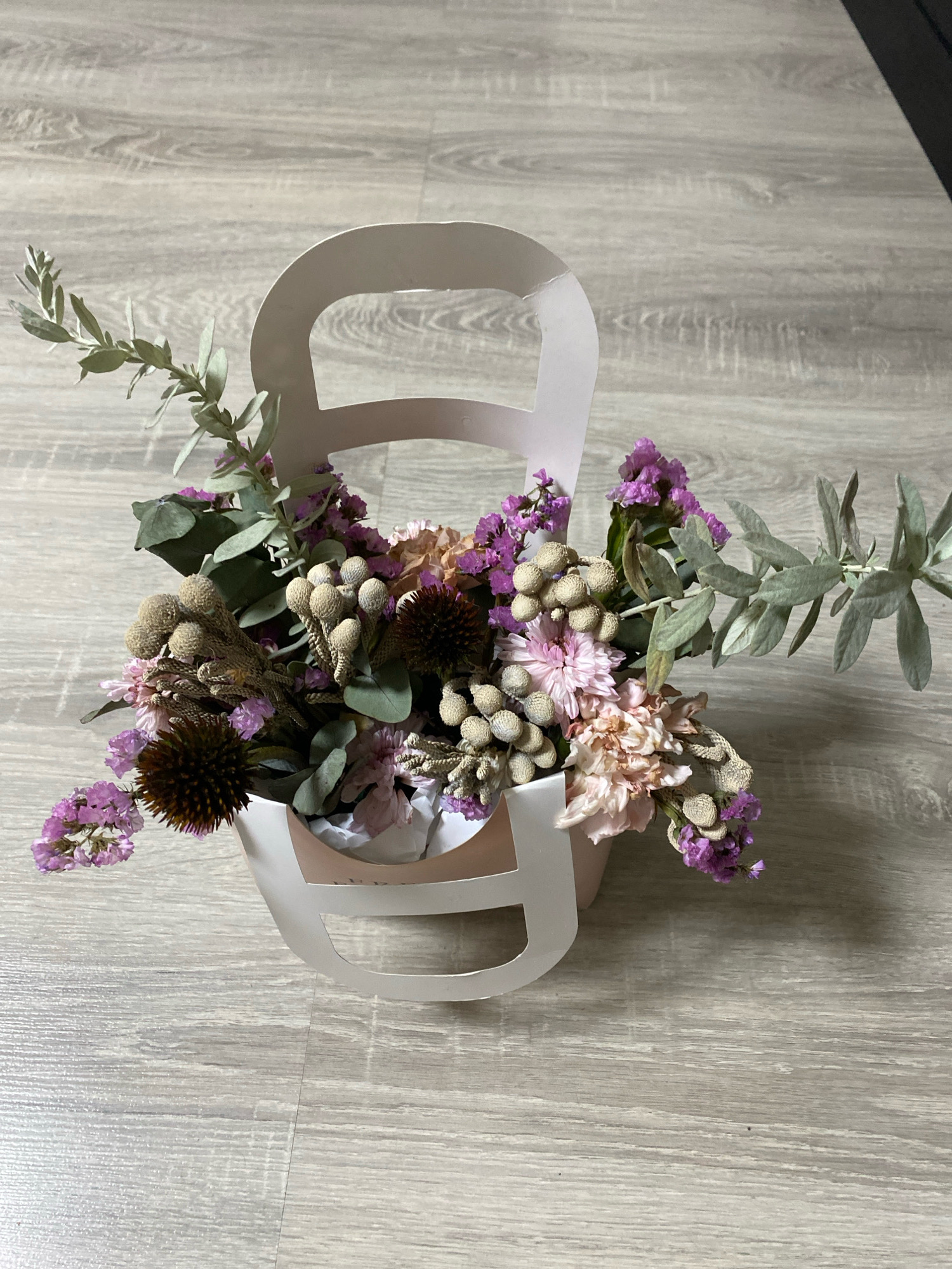 A bouquet of dried flowers is displayed in a light pink paper gift bag with cut-out handles. The bouquet features various textures and colors, including dried purple statice, light pink and beige carnation-like flowers, cone-shaped dried elements, and sprigs of grey-green eucalyptus. Two dark brown, spiky dried flower heads are prominent. The gift bag is positioned on a light grey wooden surface, appearing to be flooring. The lighting suggests an indoor environment, possibly a home or retail space. The image does not contain any people or show any specific landmarks or distinguishing features of Ivanteyevka, Russia.