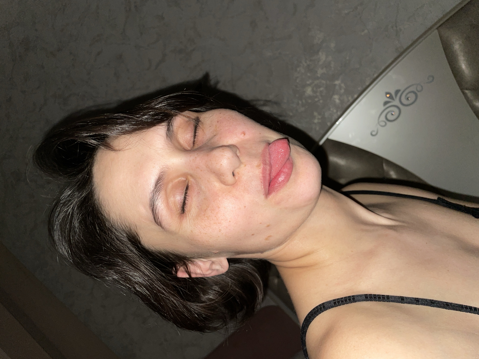 A young woman with short, dark hair is lying down with her eyes closed and tongue sticking out. She has freckles on her face. She is wearing a thin-strapped black top. The background is a textured wall. Part of a white object with decorative swirls is also visible. It is difficult to determine if this image was taken in Izhevsk, Russia, as there is no information to link it to this location.