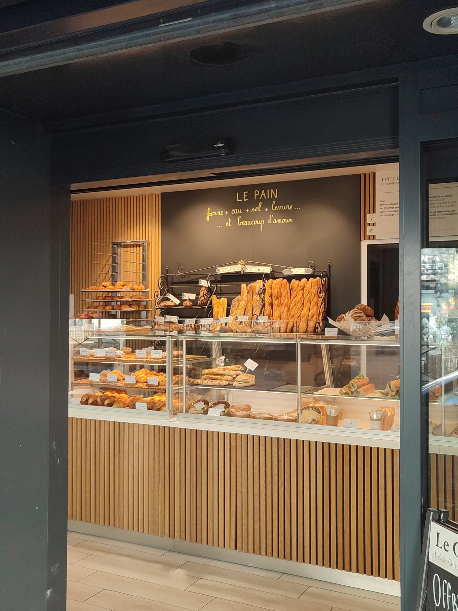 The image shows the interior of a bakery in Boulogne-Billancourt, France. The bakery features a display of various baked goods, including baguettes, croissants, and sandwiches, arranged on shelves within glass cases. A large chalkboard behind the display has text written in yellow, including "LE PAIN" and a slogan about flour, water, salt, yeast, and love. The bakery's interior is decorated with wooden paneling. To the right, a sign with "LE C" and "LES GR" is visible, partially obscured by a dark pillar. The lighting suggests it is daytime.