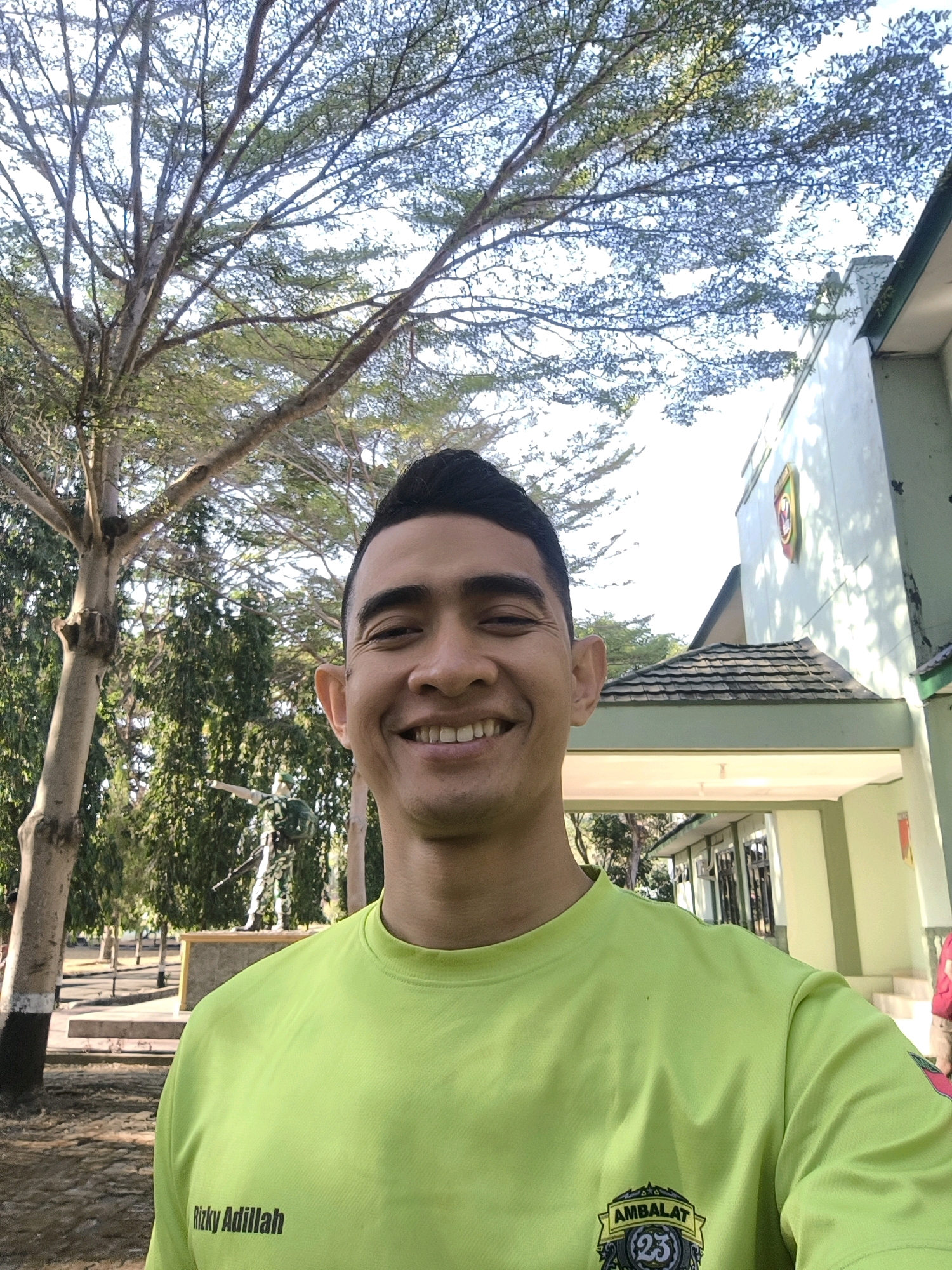 An outdoor, waist-up, medium shot captures a smiling man with dark hair wearing a neon green t-shirt. The man is positioned in the foreground, facing the camera with a slight head tilt. His t-shirt has black text that reads "Rizky Adillah" on the left side and a circular emblem with the word "AMBALAT" and the number "23" on the right. Behind the man, a light green building with a dark roof is visible, featuring a circular emblem on its facade. Trees with sparse leaves and branches are prominent in the background, with sunlight filtering through them. The ground appears to be made of dirt and gravel. In the mid-ground, a statue of a soldier in camouflage uniform holding a rifle stands on a raised platform. The overall setting suggests an outdoor area, possibly a military compound or training ground, given the soldier statue and building architecture.