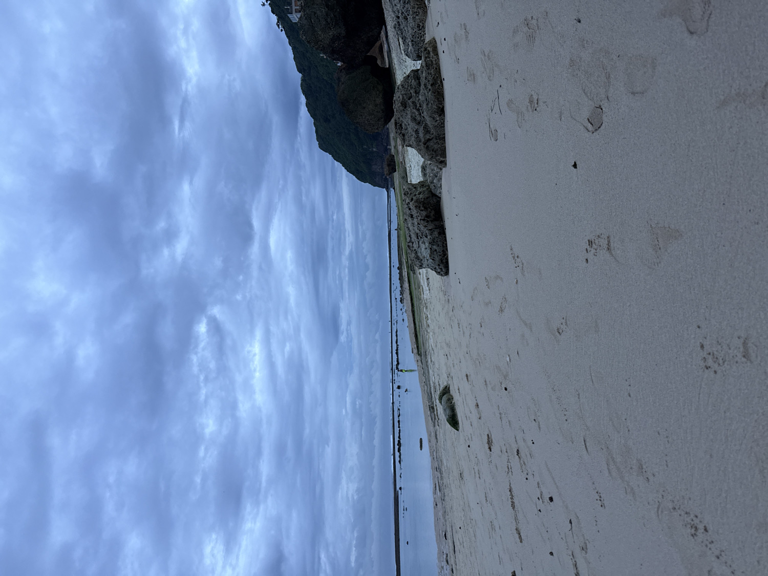 The image displays a tranquil beach scene under an overcast sky, likely in Kuta Selatan, Indonesia. The upper portion of the frame is dominated by a heavy, uniform layer of dark grey clouds, indicating subdued light and possibly a very cloudy day.
On the left side, a dark, vegetated cliff or hillside rises, with hints of structures at its peak barely visible against the clouded sky. A calm body of water, likely the ocean, stretches across the middle of the frame, its surface appearing still and dark, reflecting the muted tones of the sky. In the distant water, two very small, indistinct figures can be observed near the waterline, suggesting human presence.
The right side of the image features a wide expanse of light-colored sand, marked extensively with numerous footprints, indicating recent human activity. Several dark rocks are scattered along the sandy shore, particularly near the transition to the water and the cliff base. The overall atmosphere is one of quiet solitude, characterized by the muted colors and the heavy, continuous cloud cover. No discernible text is visible in the image.