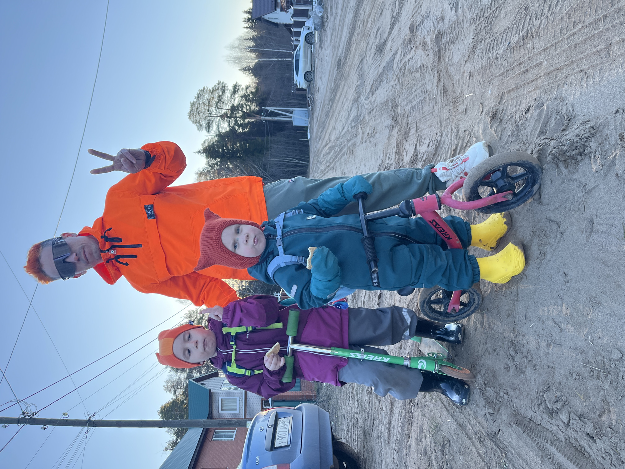 The image shows a man and two young children outdoors. The man, wearing an orange jacket and sunglasses, is making a peace sign. One child, wearing a blue jumpsuit and yellow boots, stands next to him, holding a small bike. The other child, dressed in a purple jacket and black boots, is standing with a green scooter. Both children appear to be eating. A blue Lada car is partially visible in the foreground, with a license plate number "Y 907 AH 59". The ground appears to be sandy. In the background are houses and trees. The image location is Chaikovsky, Russia, based on the license plate details of the vehicle.