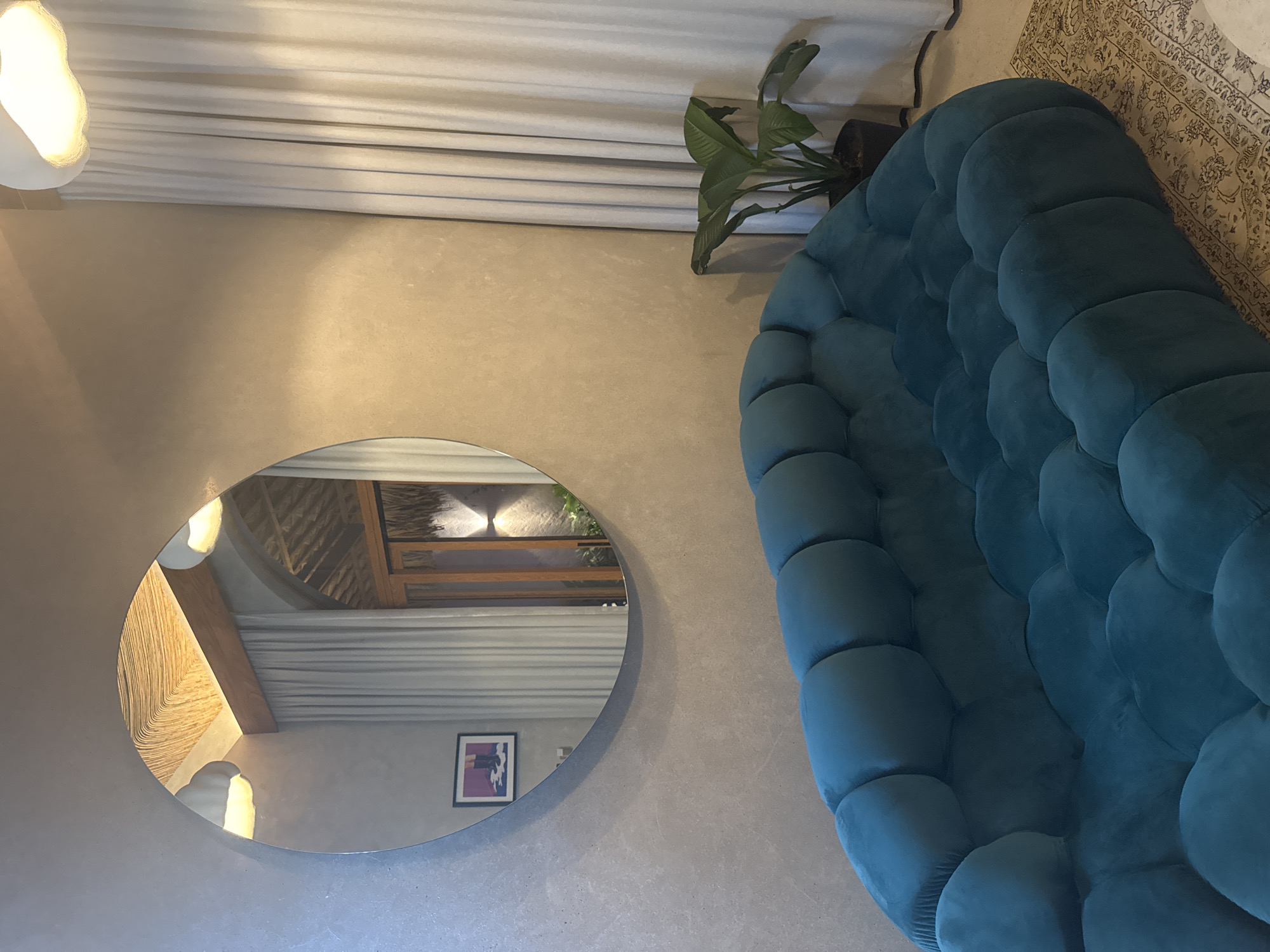 The scene depicts an interior space, consistent with a modern lounge or guest area in El Nido, Philippines. In the foreground and midground, a large, curving sofa upholstered in teal velvet features a distinctive bubble or modular tufted design. The walls are finished with a textured, light gray, concrete-like material. A large, circular, frameless mirror is mounted on the left wall above the sofa.
The mirror's reflection reveals a high ceiling section with woven natural material and a prominent wooden beam, along with two modern, white, cloud-shaped pendant light fixtures. Also visible in the reflection is a dark wood-framed doorway leading to an outdoor or semi-outdoor area featuring a thatched roof detail, a wall-mounted up-and-down light, and green foliage. A framed picture, depicting a person's lower legs in dark boots standing in a purple puddle, is reflected on an opposing wall.
To the right of the sofa, floor-to-ceiling light-colored curtains obscure a window or doorway. A potted plant with broad, dark green leaves sits on the floor between the sofa and the curtains. A patterned rug in muted tones is partially visible under the right side of the sofa. No individuals are present, and no actions or interactions are depicted.