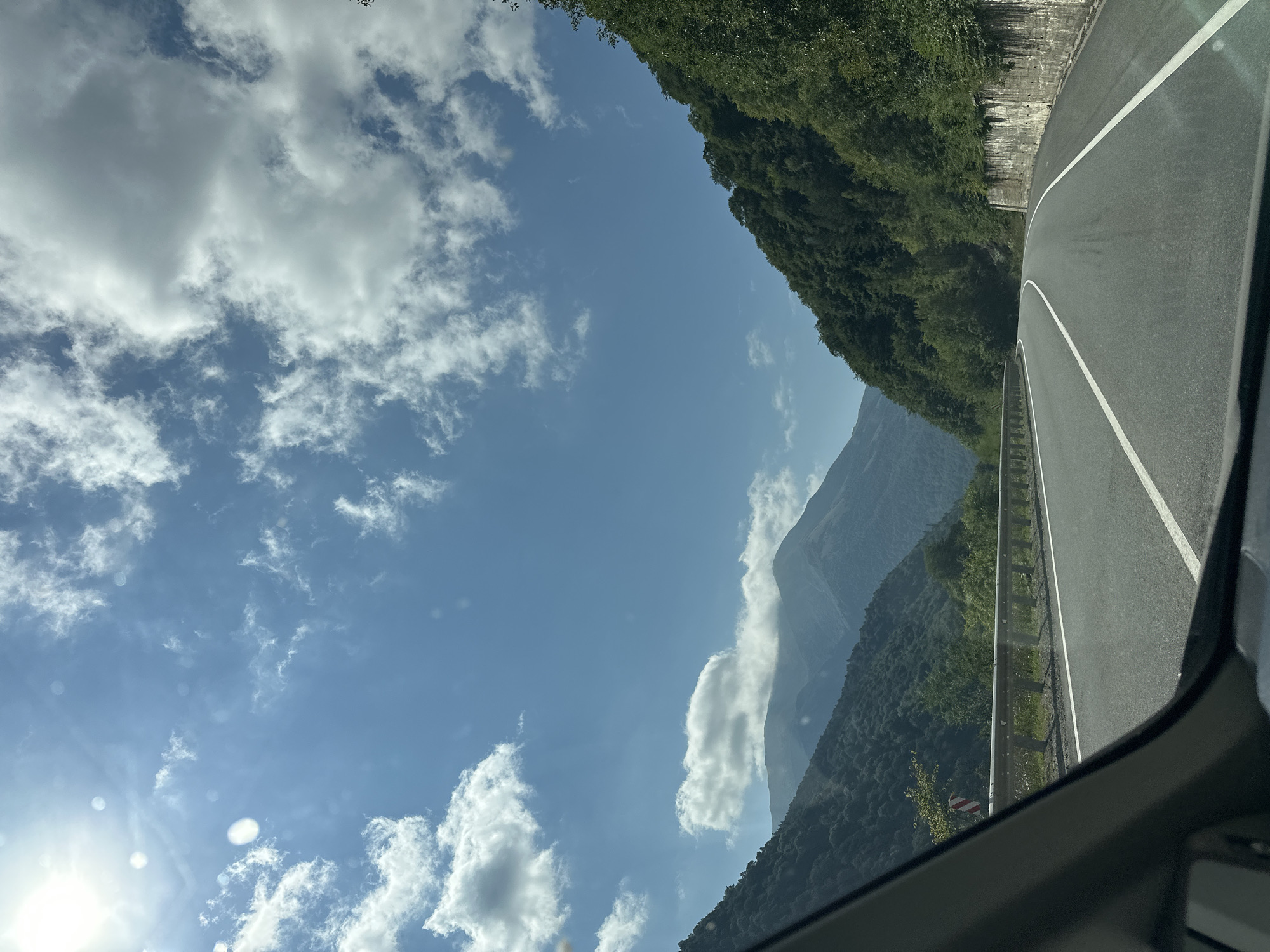 The image captures a view from inside a vehicle traveling along a paved road in the mountainous region of Nizhnii Arkhyz, Russia. The road curves gently to the right, with white lane markings visible. On the left, a metal guardrail separates the road from a steep, tree-covered slope leading up to distant mountains. A red and white striped warning sign is visible on the left side of the road. On the right side of the road, a stone retaining wall is present. The sky is blue with scattered white clouds, and the sun is shining brightly in the upper left corner, creating some lens flare. The landscape is characterized by lush green trees covering the slopes of the mountains, with some appearing hazy in the distance. The overall impression is a scenic drive through a natural, mountainous environment.