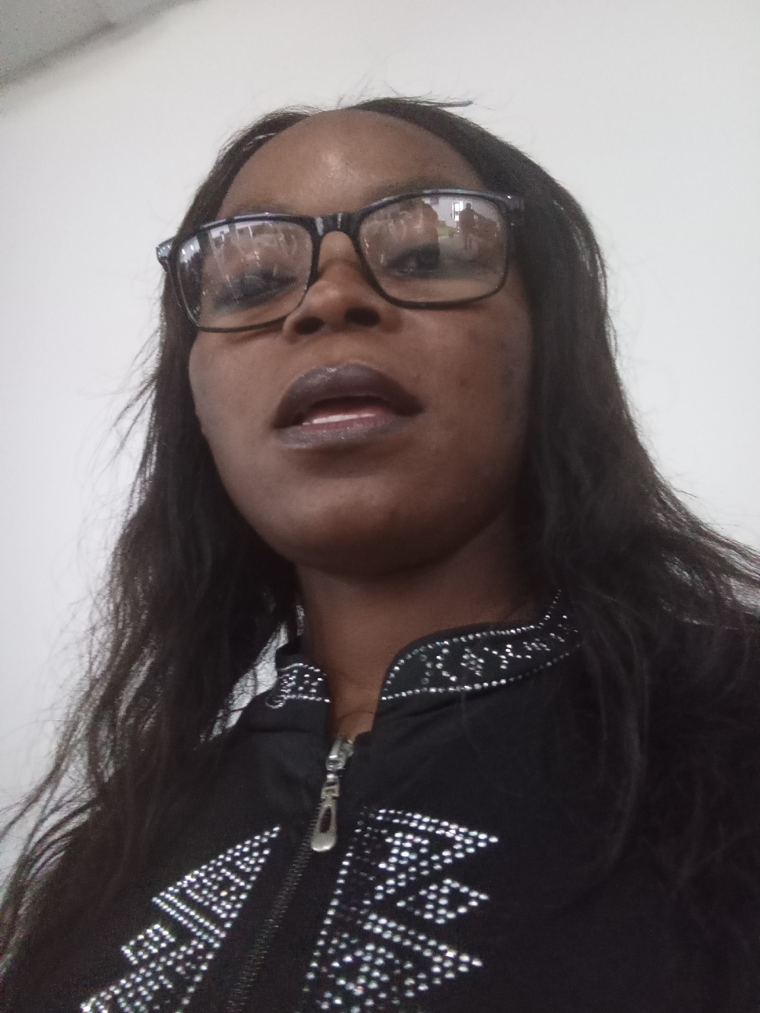 A woman is looking up into the camera. She is wearing black-rimmed glasses, dark lipstick, and a black jacket with silver beading around the collar and on the front. The jacket has a zipper closure. The background is a plain white wall. The image appears to be a selfie taken indoors. There are reflections of people in the woman's glasses. The scene does not provide any information about the specific location in the city of Kason, Nigeria.