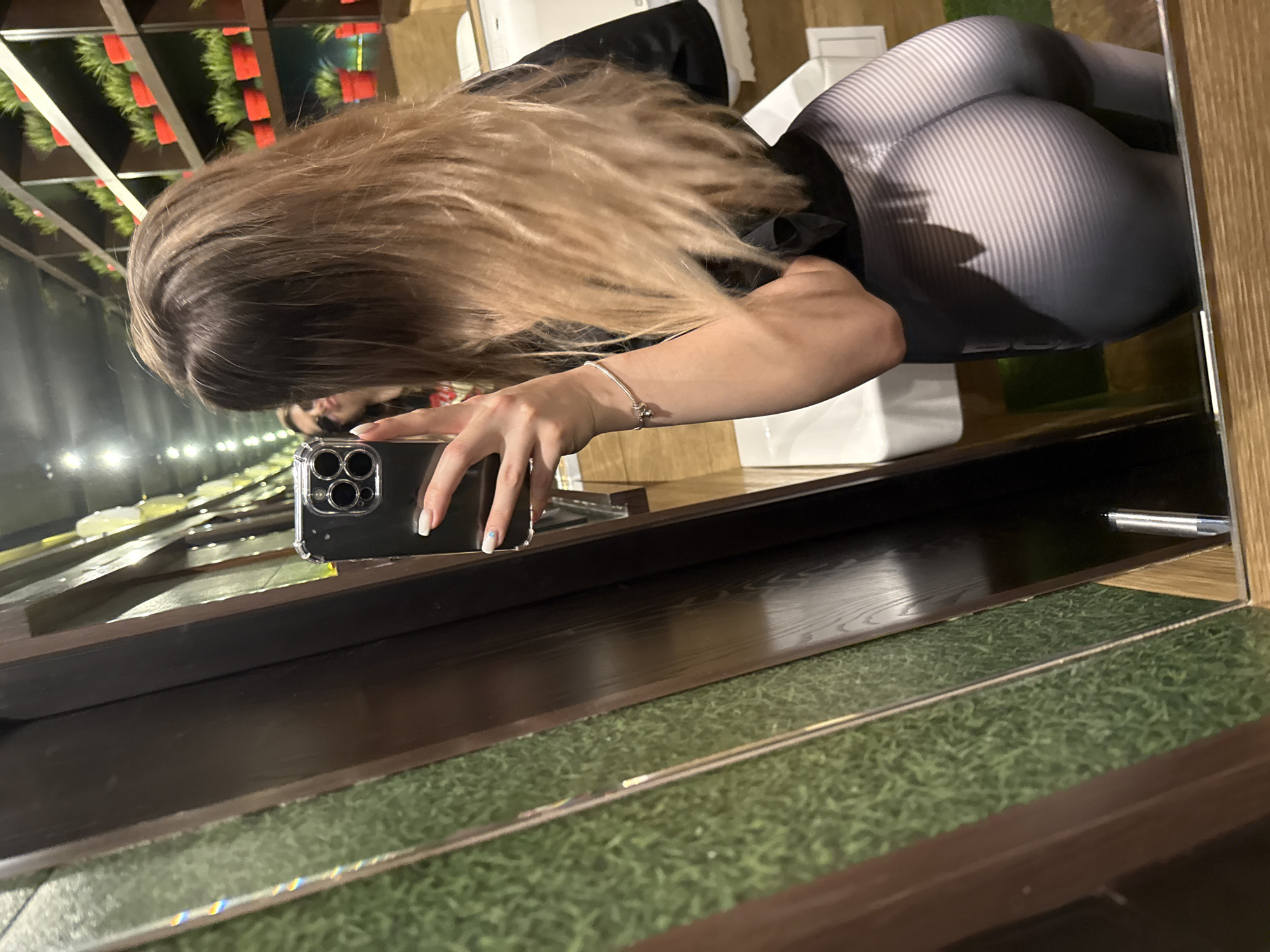 An indoor mirror selfie captures a person with long blonde and brown hair wearing a black top and grey-and-white striped leggings. The person is holding a smartphone in a clear case with their left hand, angled towards the mirror to take the photo. A silver bracelet adorns their left wrist, and their fingernails are painted white with a small blue detail on the ring finger.
The mirror reflects the person's upper body and the surrounding environment. In the background, visible in the reflection, is a ceiling structure with white beams from which green foliage and red decorative elements are suspended. Other reflected objects include white rectangular surfaces and dark wooden counters or shelves. Below the main mirror frame, a green, artificial-grass-like texture is visible on a lower surface, suggesting a decorative floor or shelf. The overall image is tilted, giving a dynamic perspective. There is no discernible text visible in the image.