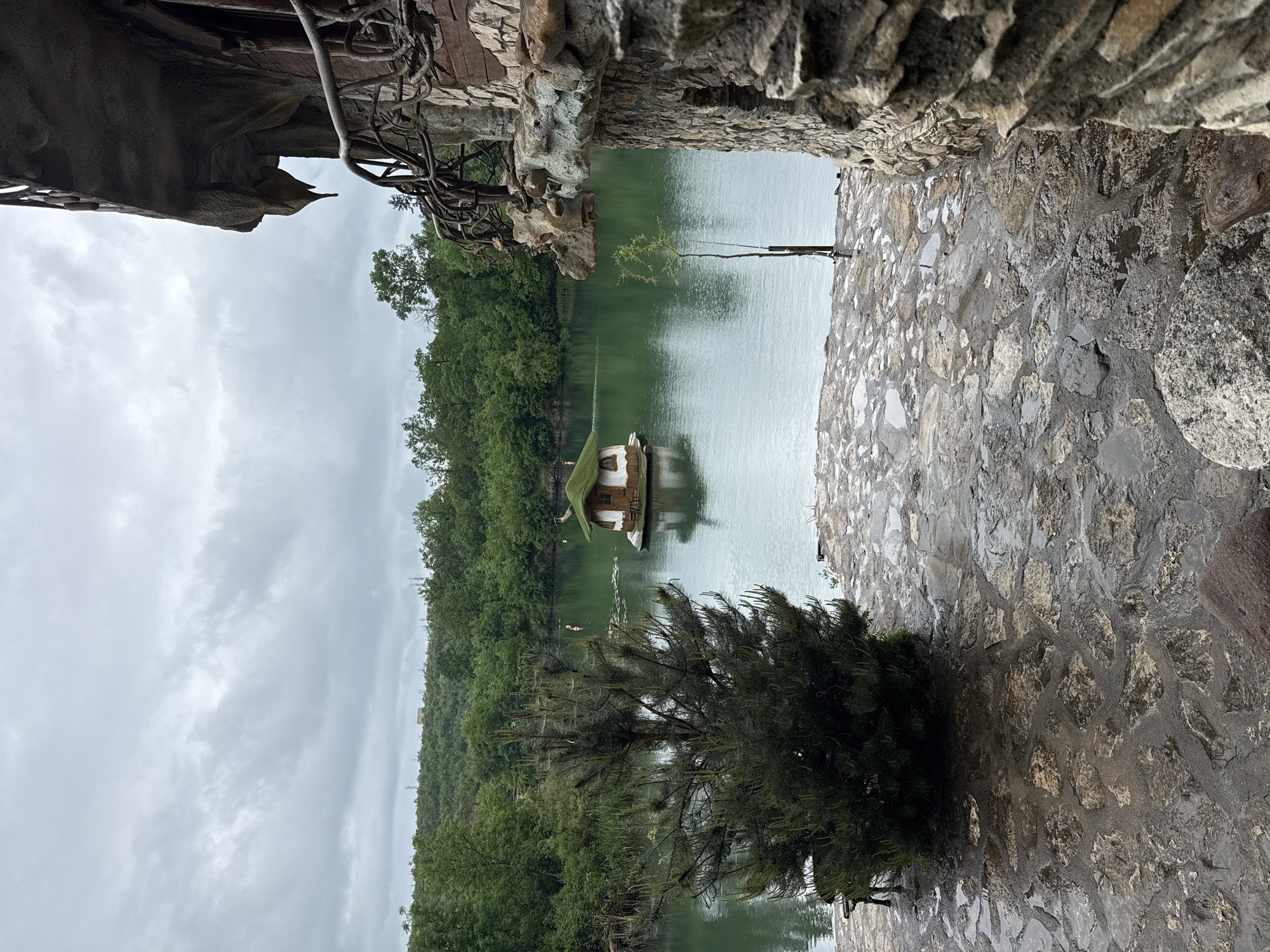 The image shows a view from a stone structure, likely part of a building or wall, overlooking a lake or pond in the city of Lukovit, Bulgaria. In the center of the water is a small, elevated wooden structure with a green roof, possibly a gazebo or small house. Dense green trees and vegetation surround the water, suggesting a natural or park-like setting. The sky is overcast and grey. The stone structure is constructed of various sized stones. Part of an overhanging stone or wooden feature is visible at the top left of the frame.