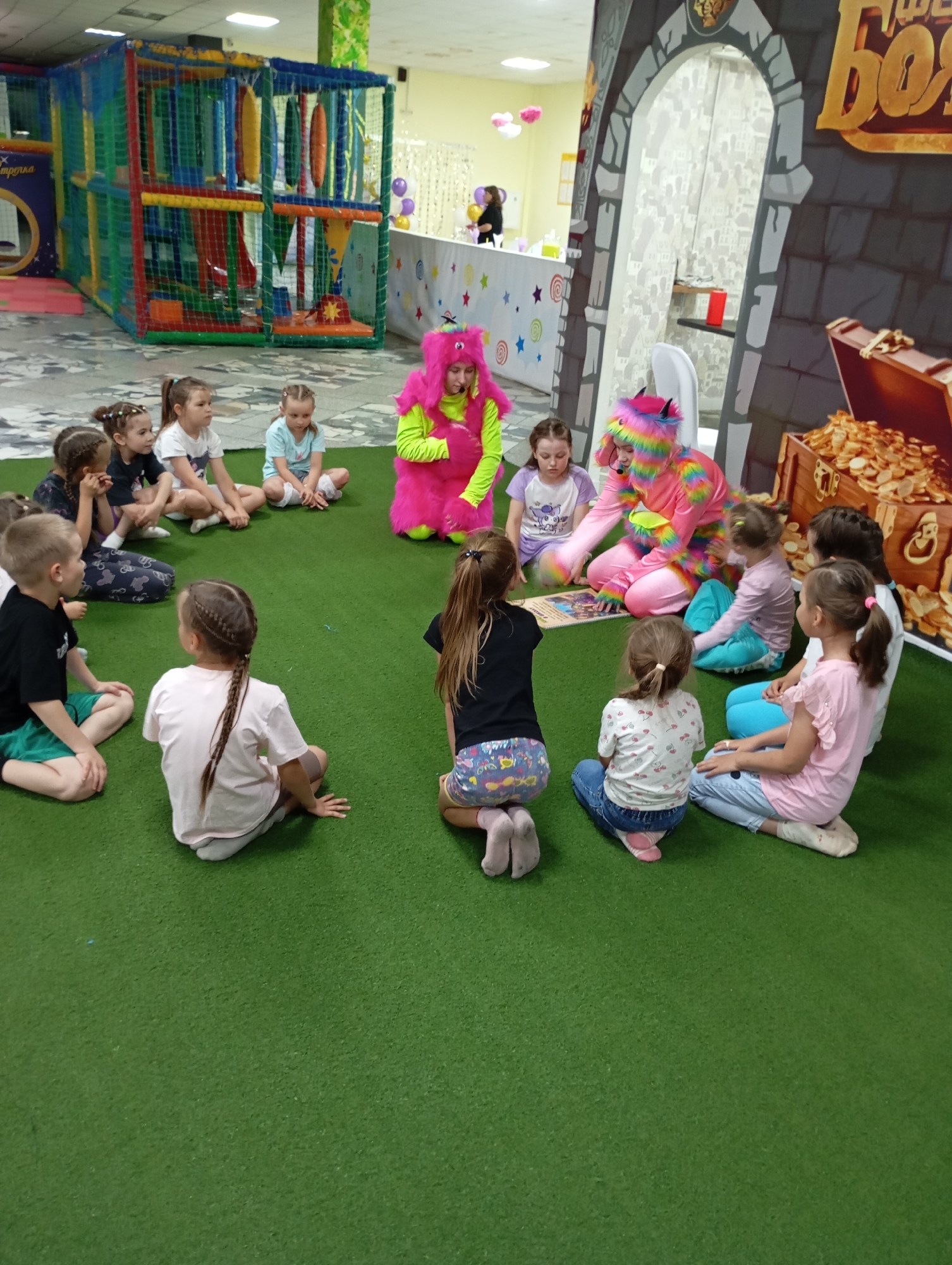 A medium, eye-level shot indoors shows a group of children seated on a green floor, gathered around two adults dressed in colorful costumes. The adults appear to be reading or presenting something to the children.
The children are of various ages, ranging from approximately 4 to 8 years old. They are dressed in casual clothing, and their expressions are attentive.
The adults are dressed in brightly colored costumes. One is in a bright pink and the other in bright pink with rainbow accents. They are positioned in the center of the group, interacting with the children.
The setting appears to be a play area or a child-friendly environment. In the background, there is a multi-level play structure with netting and colorful tubes. There is also a backdrop depicting a castle entrance and a treasure chest filled with gold coins.