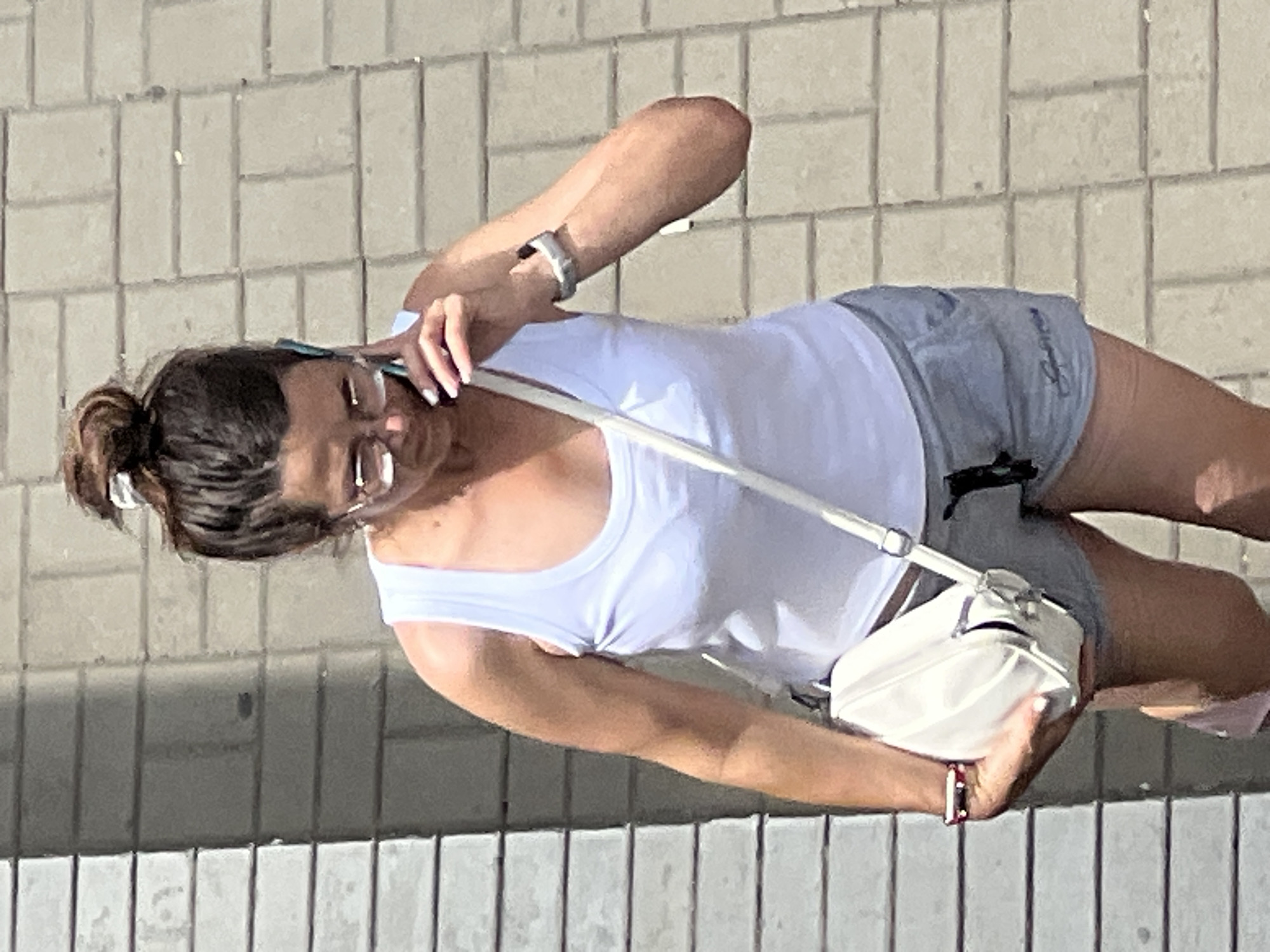 A woman with dark hair pulled into a bun on top of her head, wearing glasses, a white tank top, and grey shorts, is walking on a paved sidewalk in Krasnodar, Russia. She is holding a light-colored smartphone to her right ear with her right hand, and her left hand is resting on a white shoulder bag. She is also wearing a watch on her left wrist and a bracelet on her left arm. The background consists of a brick wall on the left and the paved sidewalk. The lighting suggests it is daytime.