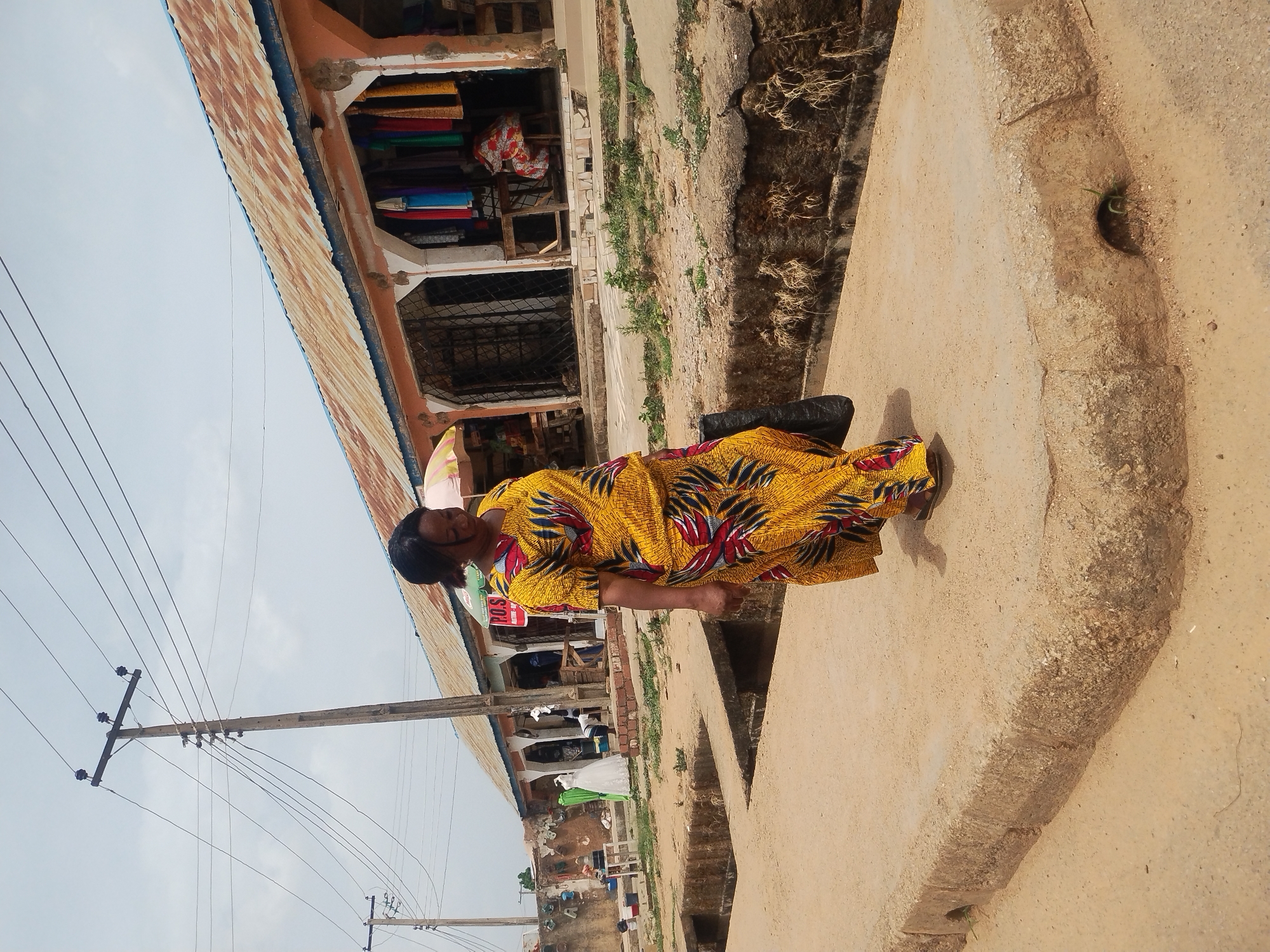 The image shows a woman standing on a concrete platform in Jos, Nigeria. She is wearing a yellow dress with red and black patterns. Behind her is a building with a rusty corrugated iron roof and an open storefront displaying various fabrics. To the left of the building are power lines and poles. The ground around the platform appears to be dry and dusty with some vegetation. The building appears to be near a busy marketplace.