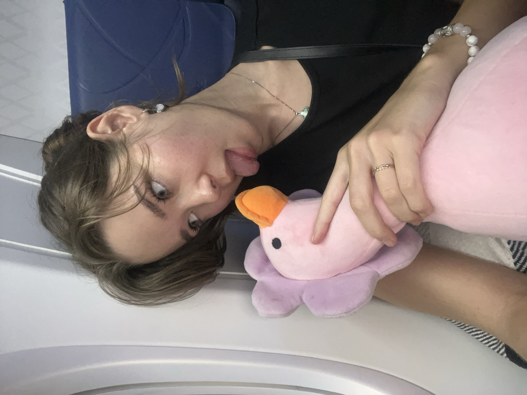 This close-up, horizontally oriented image captures a playful moment with a young woman inside what appears to be an airplane cabin, contextually located in Bac Cam Ranh Ward, Vietnam.
The subject is a young woman, seen from the chest up, with her head tilted to the side. She has light-colored eyes and brown hair, with some strands loosely framing her face. She is making a whimsical expression, sticking her tongue out towards a pink and purple plush toy. Her right hand gently cradles the plush toy.
The plush toy is a prominent object, appearing to be a stylized bird or duck with an orange beak, small black eyes, and a soft pink body, resting on a larger purple flower-like base. The woman is wearing a black top or dress, a delicate silver necklace with a light-colored heart-shaped pendant, a beaded bracelet on her right wrist, and a ring with an intricate design (possibly an infinity symbol) on her right ring finger.
The setting is an airplane interior, indicated by the blue seat visible in the background and the white cabin wall and window structure. A striped blanket or fabric is visible beneath her leg, suggesting a relaxed, possibly reclined, posture during travel. The activity depicted is a lighthearted interaction between the woman and her plush toy. No text is visible in the image. The overall mood is casual and intimate.