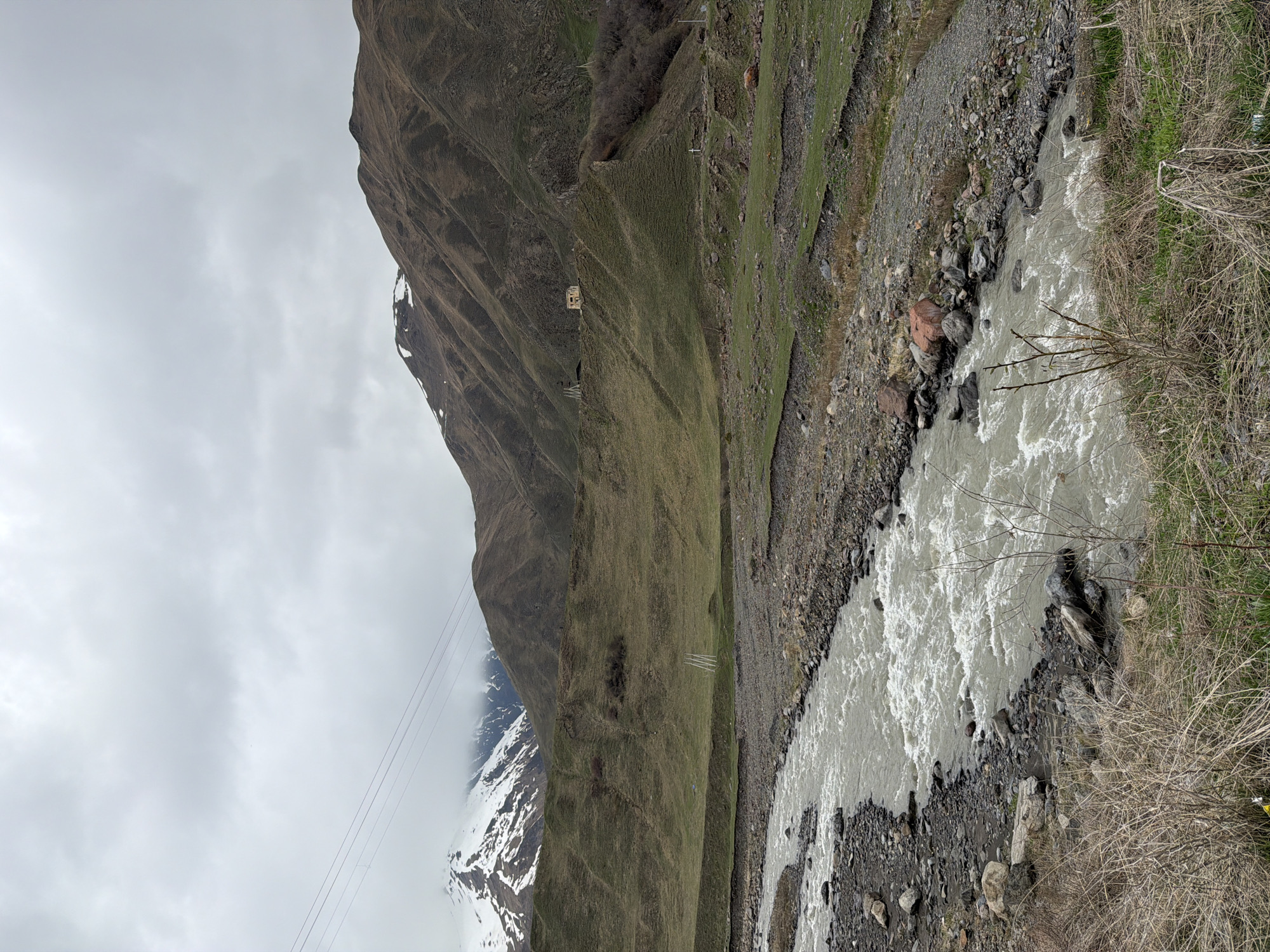 The image depicts a mountainous landscape in Khurtisi, Georgia. A river flows through a rocky riverbed in the foreground. A mountain rises to the left, partially covered in snow and cloud. A cable car system is visible, with a station and cables extending up the mountainside. Vegetation covers the mountain slopes and the riverbank.