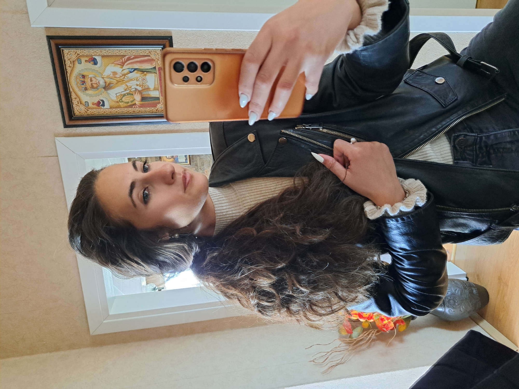 A female is taking a selfie in what appears to be a hallway or interior space. She is holding a smartphone, which obscures part of her face. She is wearing a black leather jacket over a light-colored top and has long, dark hair. Her fingernails are painted with a light color. On the wall behind her, there is framed art, possibly a religious icon. The environment seems to be a residential setting. The city and specific location are unknown.