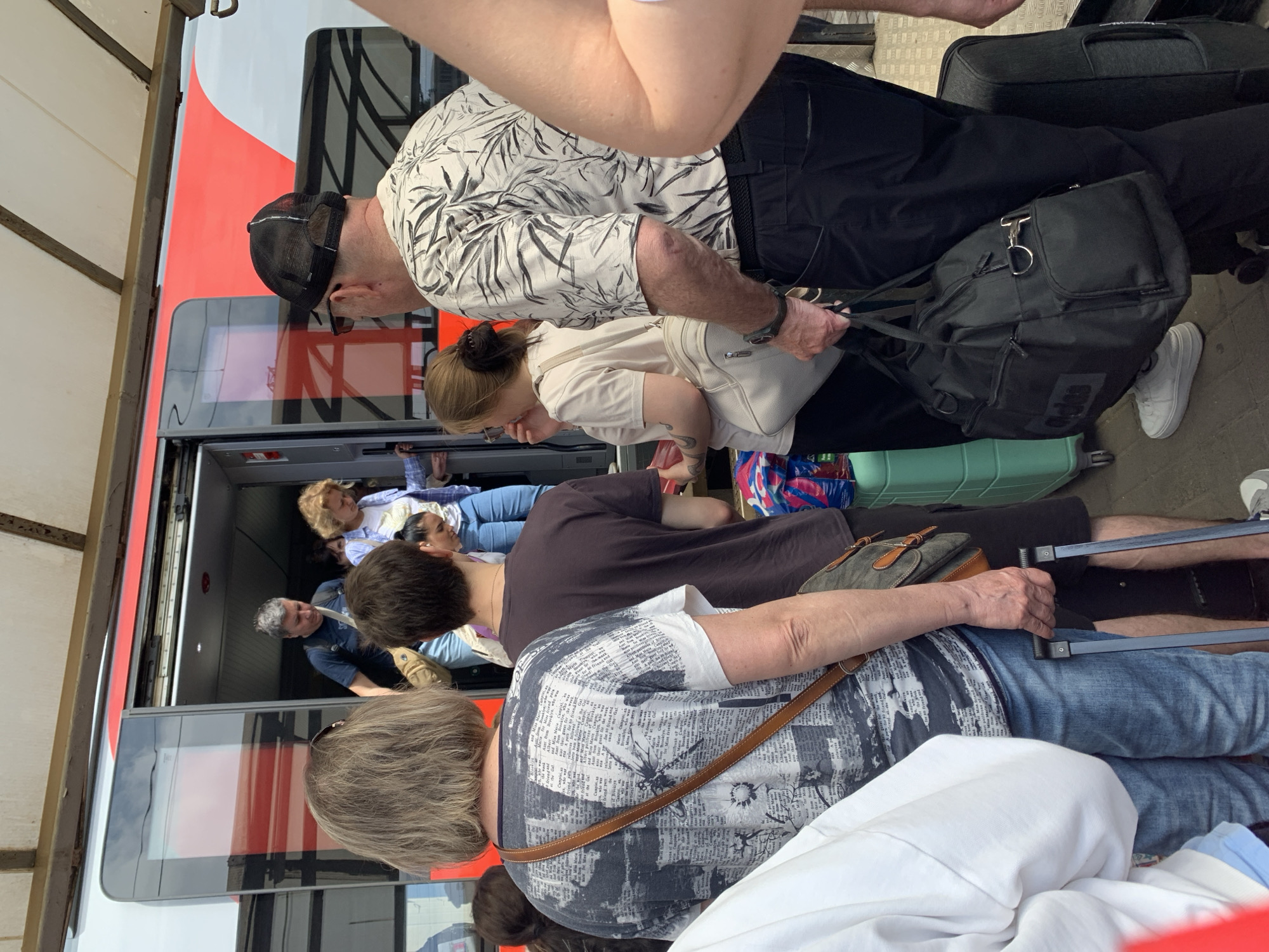 A group of people are seen entering a train, possibly at a station in an unknown city. The train is red and white, with its doors open. Individuals are carrying luggage, including rolling suitcases and handbags. Several people are already inside the train. The surrounding environment appears to be a train platform or similar boarding area.