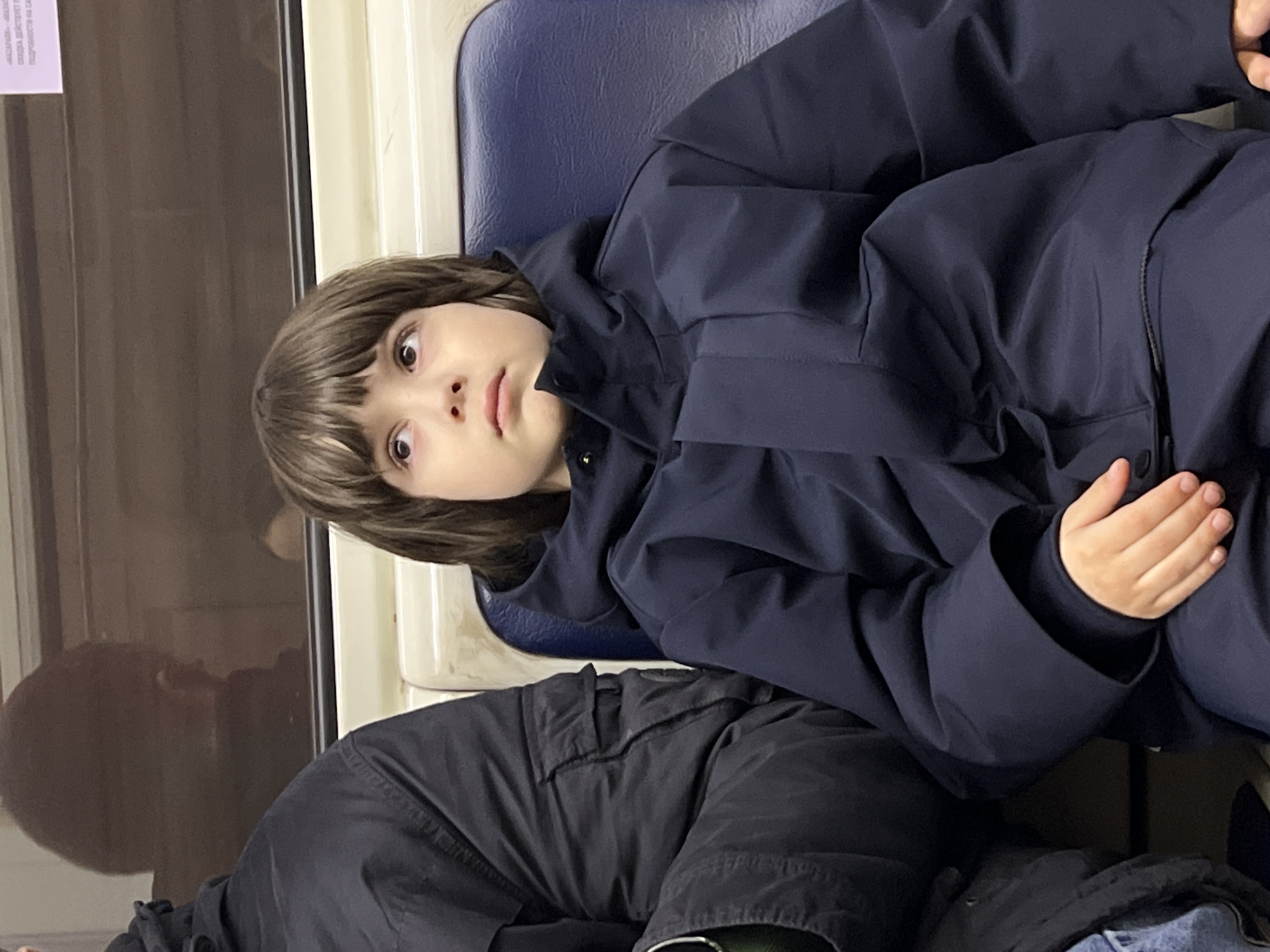 A child with brown hair and brown eyes sits on a blue seat next to a window in a public transport vehicle, likely a metro train. The child is wearing a dark blue hooded jacket and dark pants. The background indicates an urban environment, with reflections visible in the train window. It is suggested to be located in Saint Petersburg, Russia.
