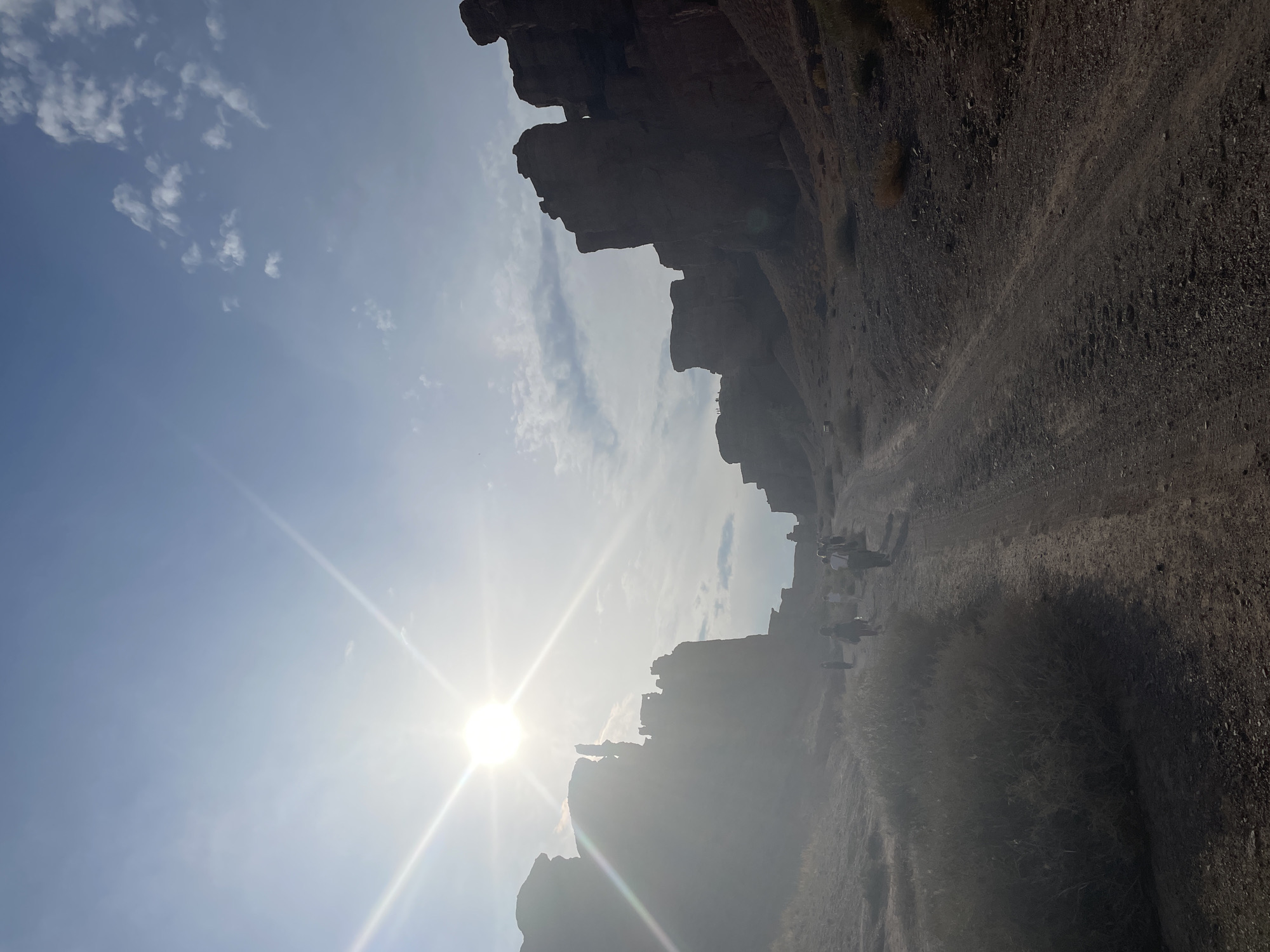 The image captures a group of people walking along a dusty path in a canyon under a bright, sunny sky. The sun, positioned in the upper left quadrant, creates a strong lens flare and illuminates the scene with a bright, hazy light. The canyon walls are tall, rugged, and appear to be made of reddish-brown rock. Vegetation is sparse, consisting mainly of dry bushes and sparse grasses. The path itself is unpaved and appears to be a dirt road with tire tracks. The people are walking in a loose group, some facing towards the camera and others away, suggesting they are on a hike or excursion. The overall setting is arid and appears to be a natural landscape, consistent with the Charyn Canyon National Park near Tamerlik, Kazakhstan.