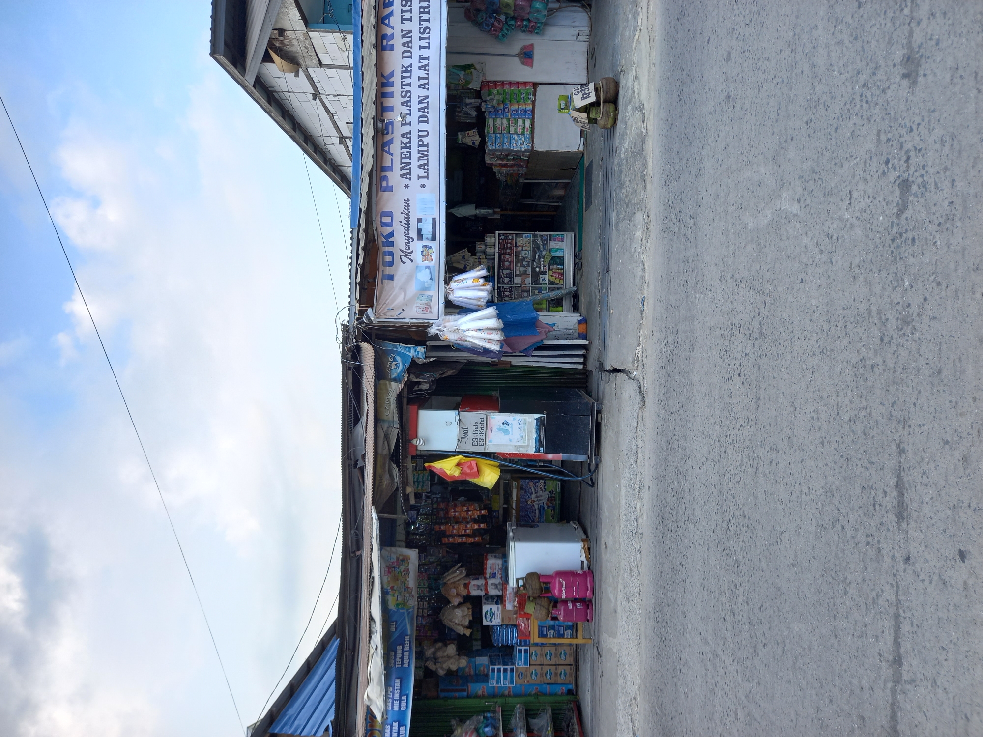 The image shows a street view in Samarinda, Indonesia. The scene features a row of small shops with corrugated metal roofs and awnings. The shops are selling various items, including plastic goods, food products, and possibly cooking gas cylinders. One shop has a sign that reads "TOKO PLASTIK RAFA" and advertises "ANEKA PLASTIK DAN TISU" and "LAMPU DAN ALAT LISTRIK." Another shop has a sign listing various food items like "GAS LPG," "MIE INSTAN," "TEPUNG," and "GULA." In front of the shops, there is a paved road and sidewalk. A few items, such as gas cylinders and baskets, are placed outside the shops. The sky above is partly cloudy. The overall impression is of a busy marketplace or commercial street in a residential area.